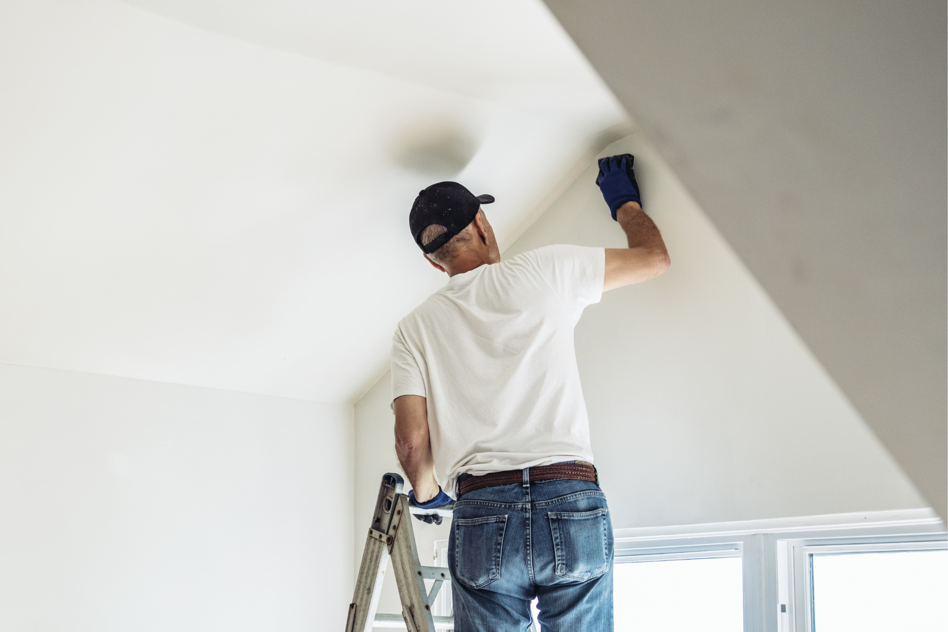 Person painting a white ceiling while standing on a ladder in a bright room