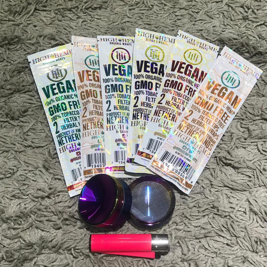 Five vegan gummies packets, two purple containers, and a pink-and-silver lighter on a gray rug