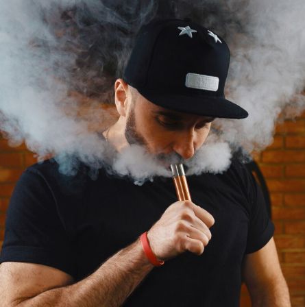 Man in black cap and shirt exhaling vapor through a vape device, surrounded by thick smoke