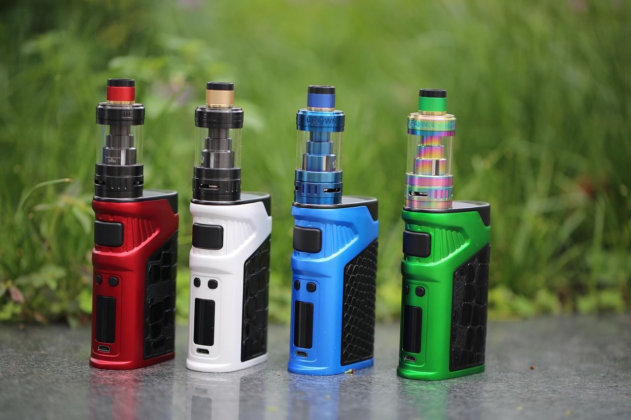 Four colorful vaping devices—red, white, blue, and green—standing on a wet surface outdoors.