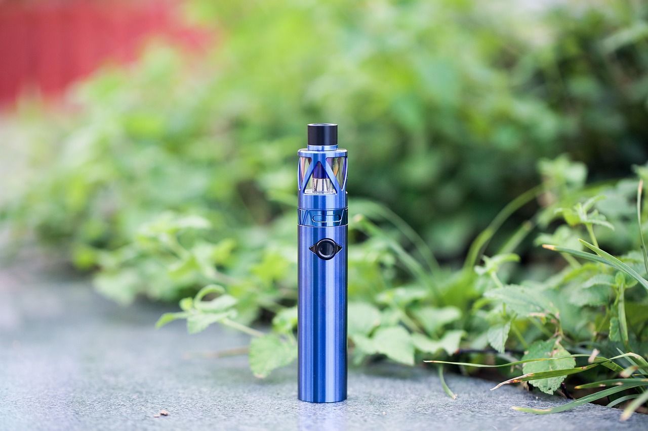 Blue vape pen standing on a pavement with blurred green foliage in the background