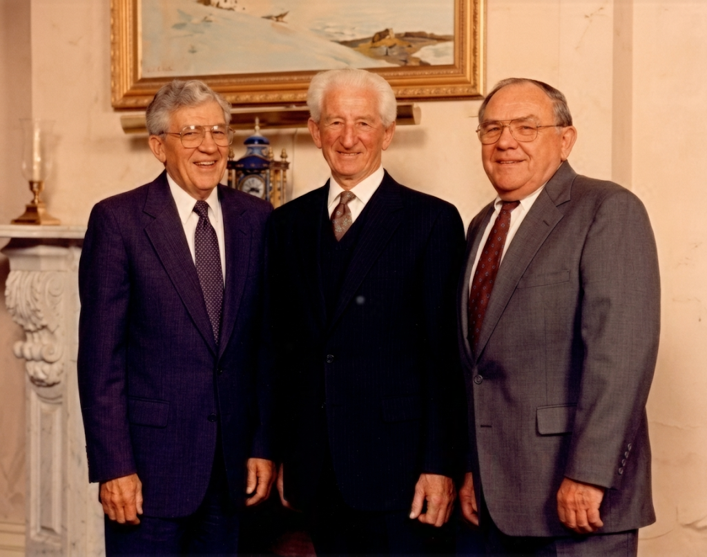 Three men in suits smiling indoors in front of a framed painting and a decorative vase.