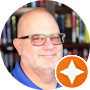 A bald man wearing glasses and a blue shirt is standing in front of a bookshelf.