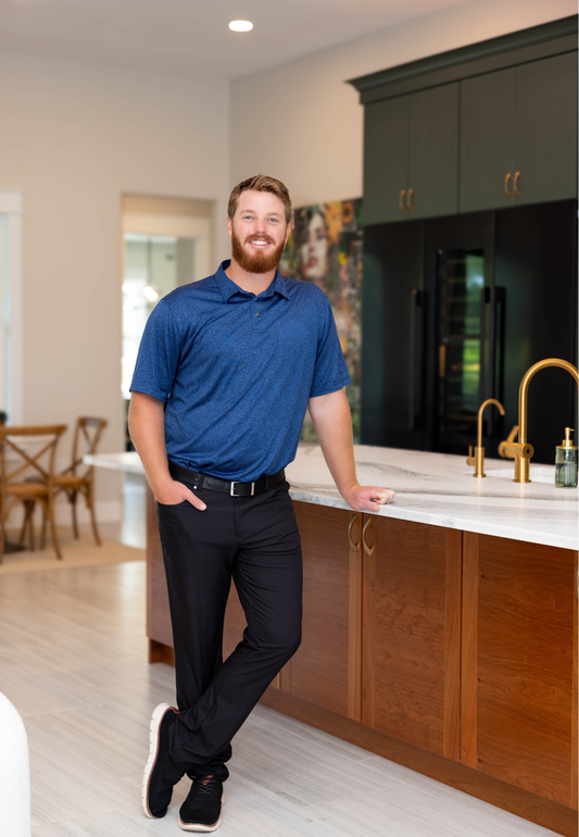 Man leaning on a kitchen island in a modern kitchen, wearing a blue shirt and black pants.
