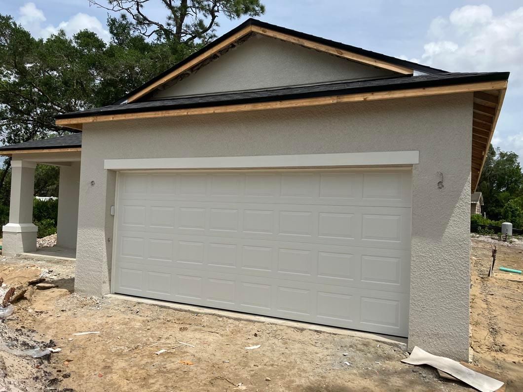 Detached garage with a gray door and beige siding, shown under construction with dirt in front