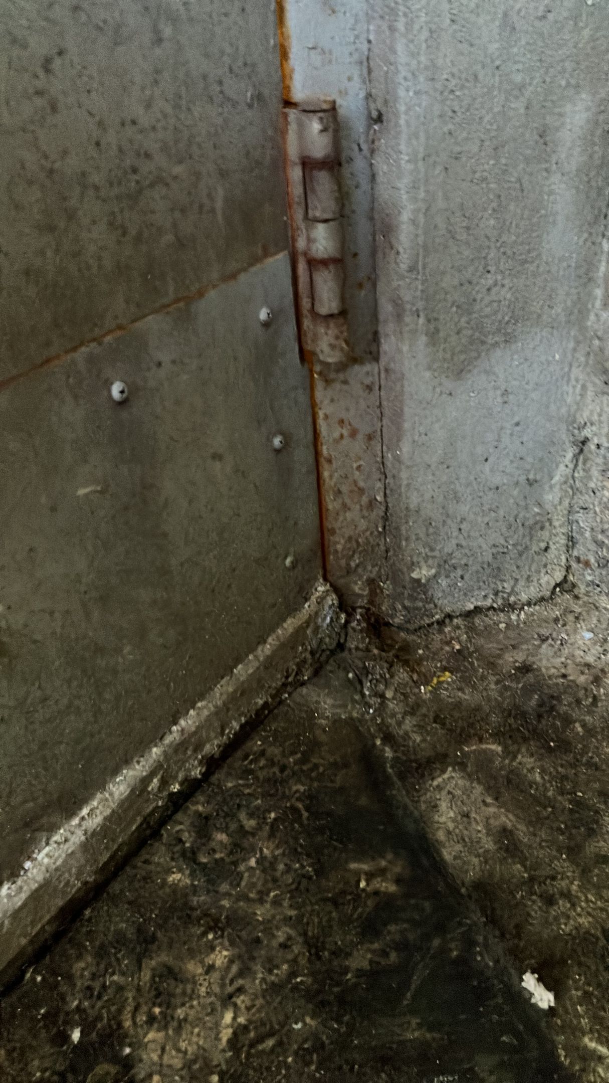 Rusty metal hinge on a grimy concrete wall in a dirty corner with stained floor.