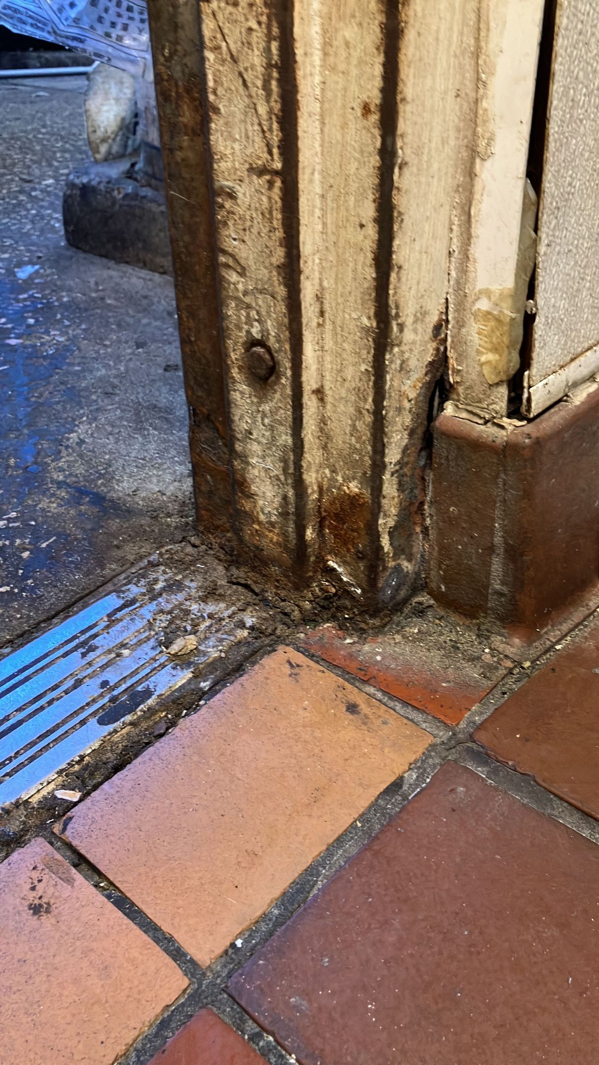 Close-up of a worn wooden door frame with rust stains beside wet terracotta floor tiles.
