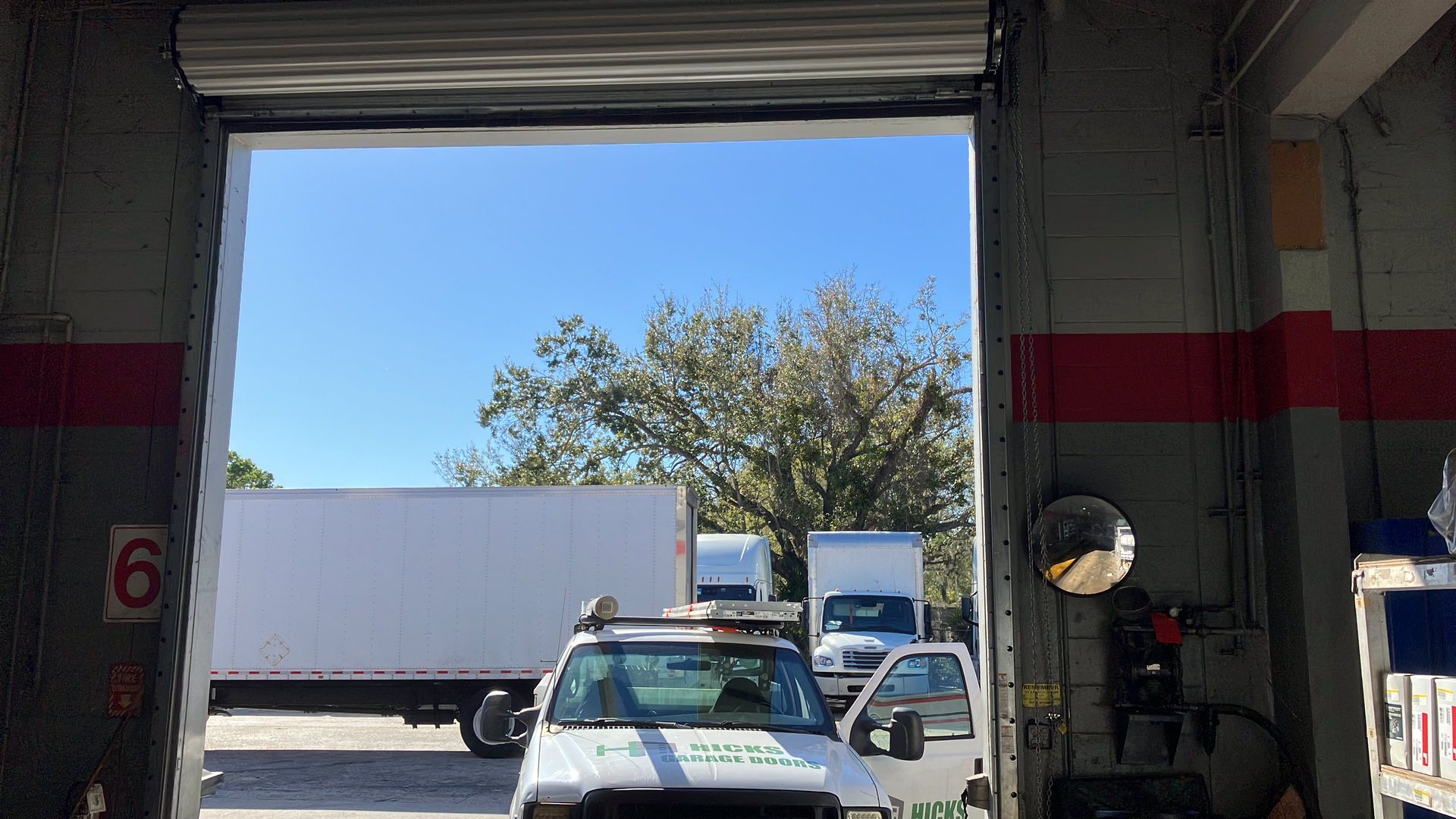 View from a garage bay to a sunlit street with parked trucks and a tree beyond the doorway