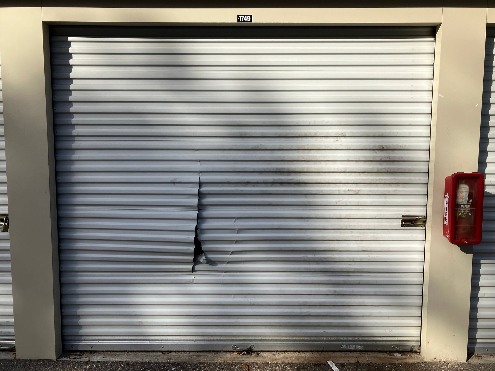 Closed white metal roll-up door with a dark damaged tear in the center and a red fire extinguisher on the right