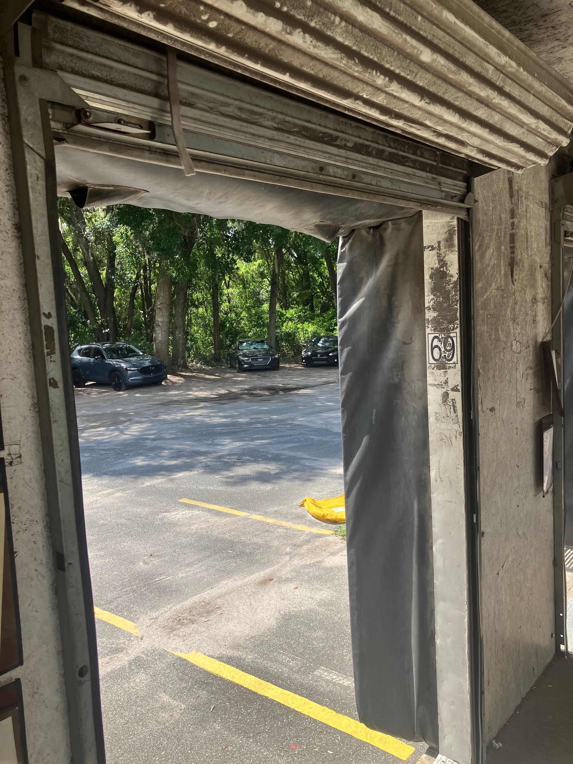 View from a garage doorway to a sunny parking lot and trees, with a yellow parking line and a dark hanging curtain.