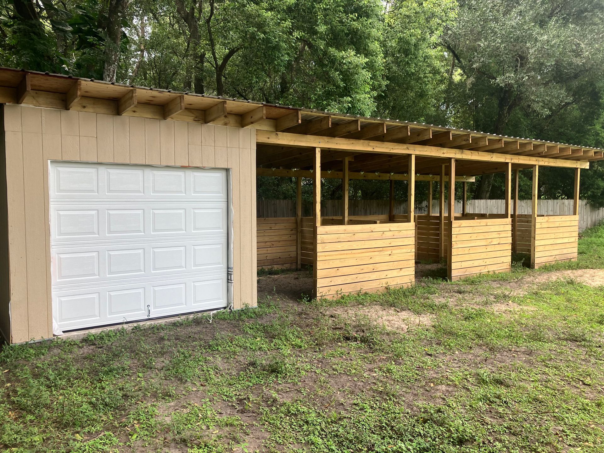Rural shed with a white garage door and open wooden stalls under a slanted roof, surrounded by trees.