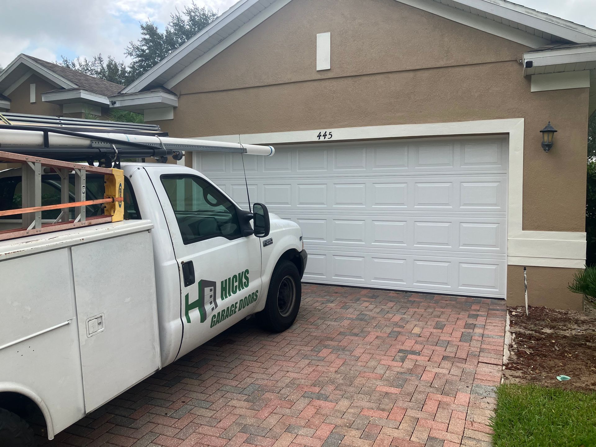 White service truck parked in a driveway in front of a beige garage door, with a ladder rack on top.