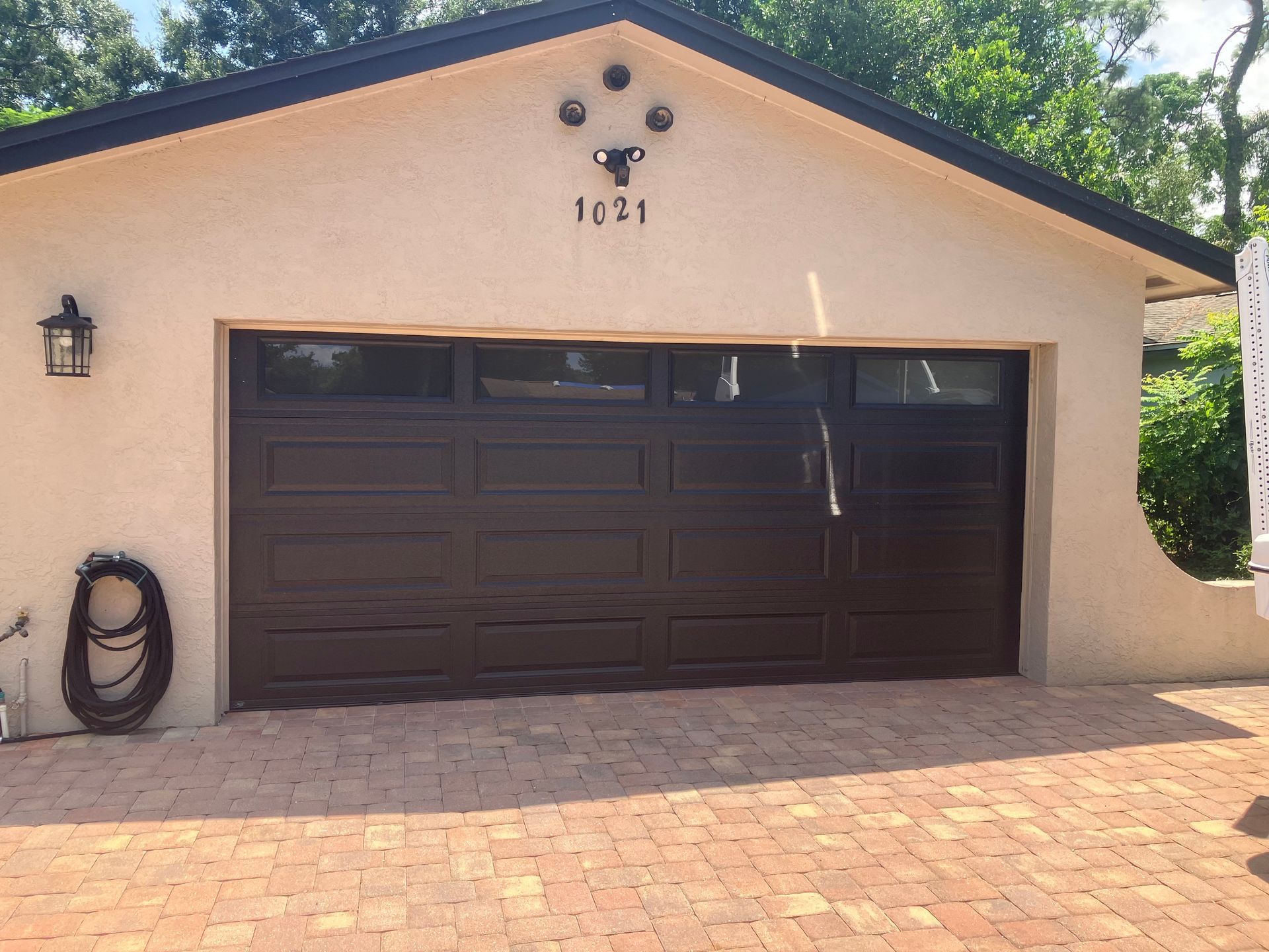 Single-story house with beige stucco, dark garage door, and brick driveway under trees