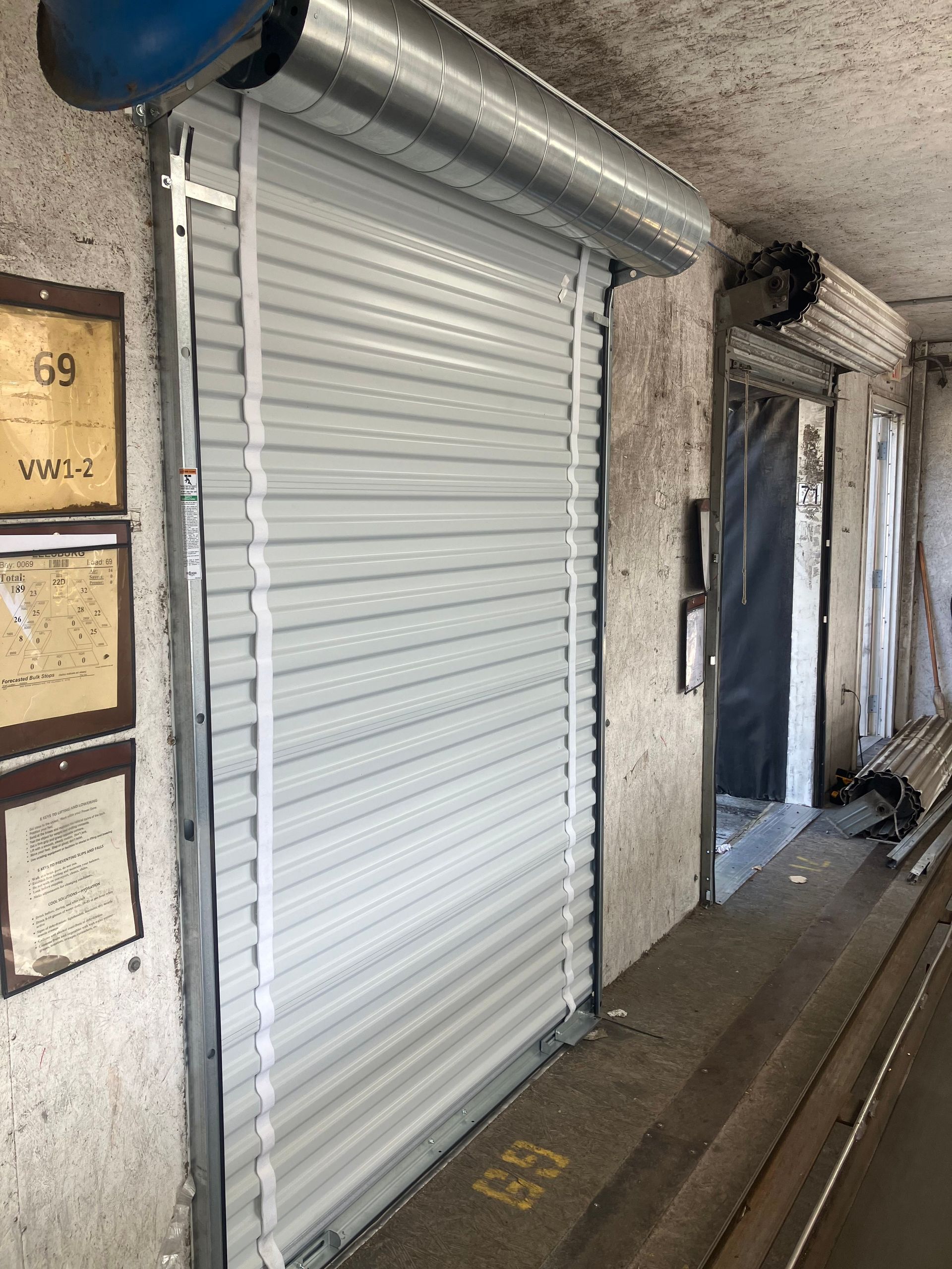 Closed white metal storefront shutter in a narrow covered walkway with posted notices on the left wall.