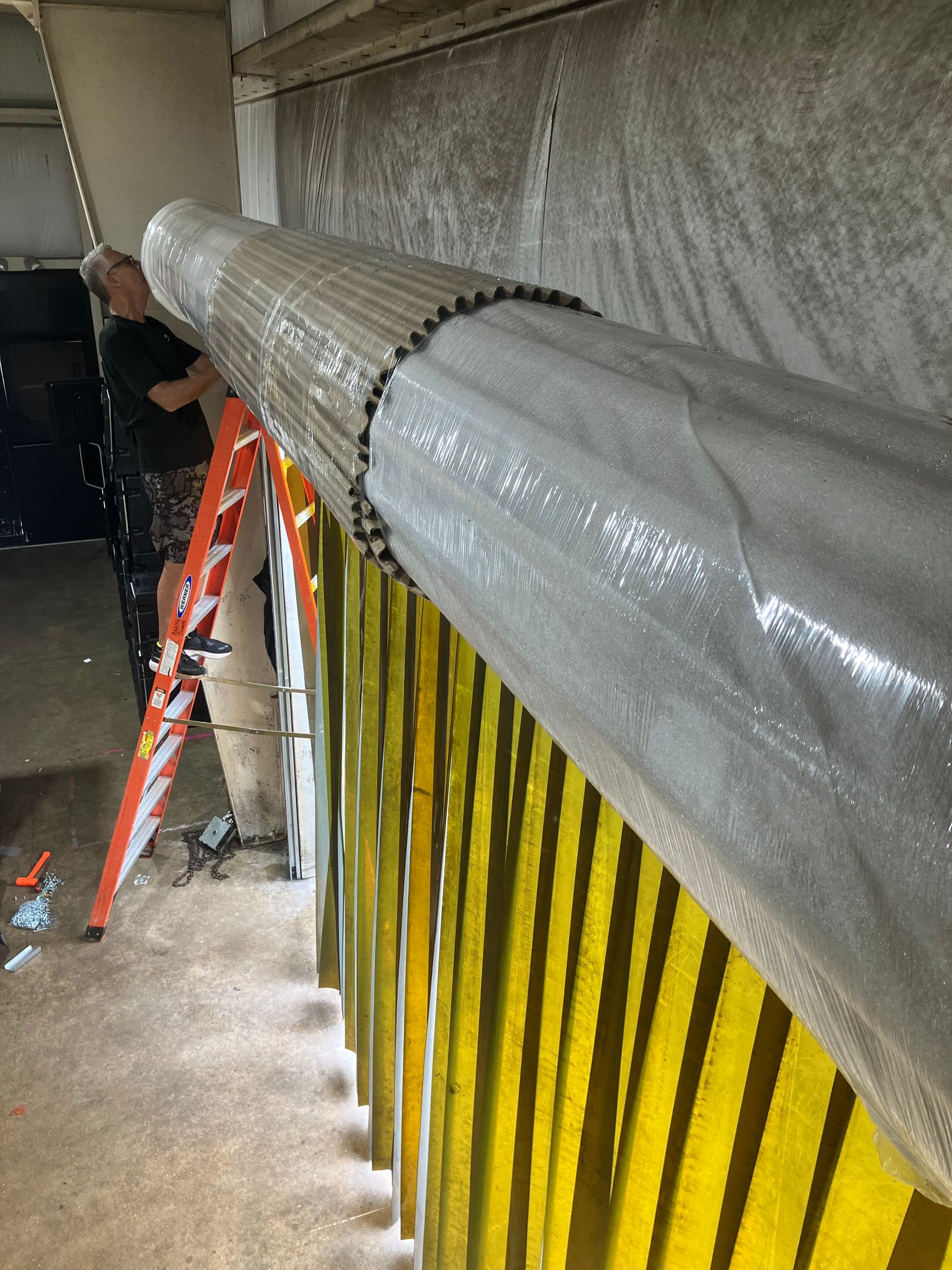 Worker wrapping a large duct with silver insulation beside a yellow railing and ladder in a stairwell