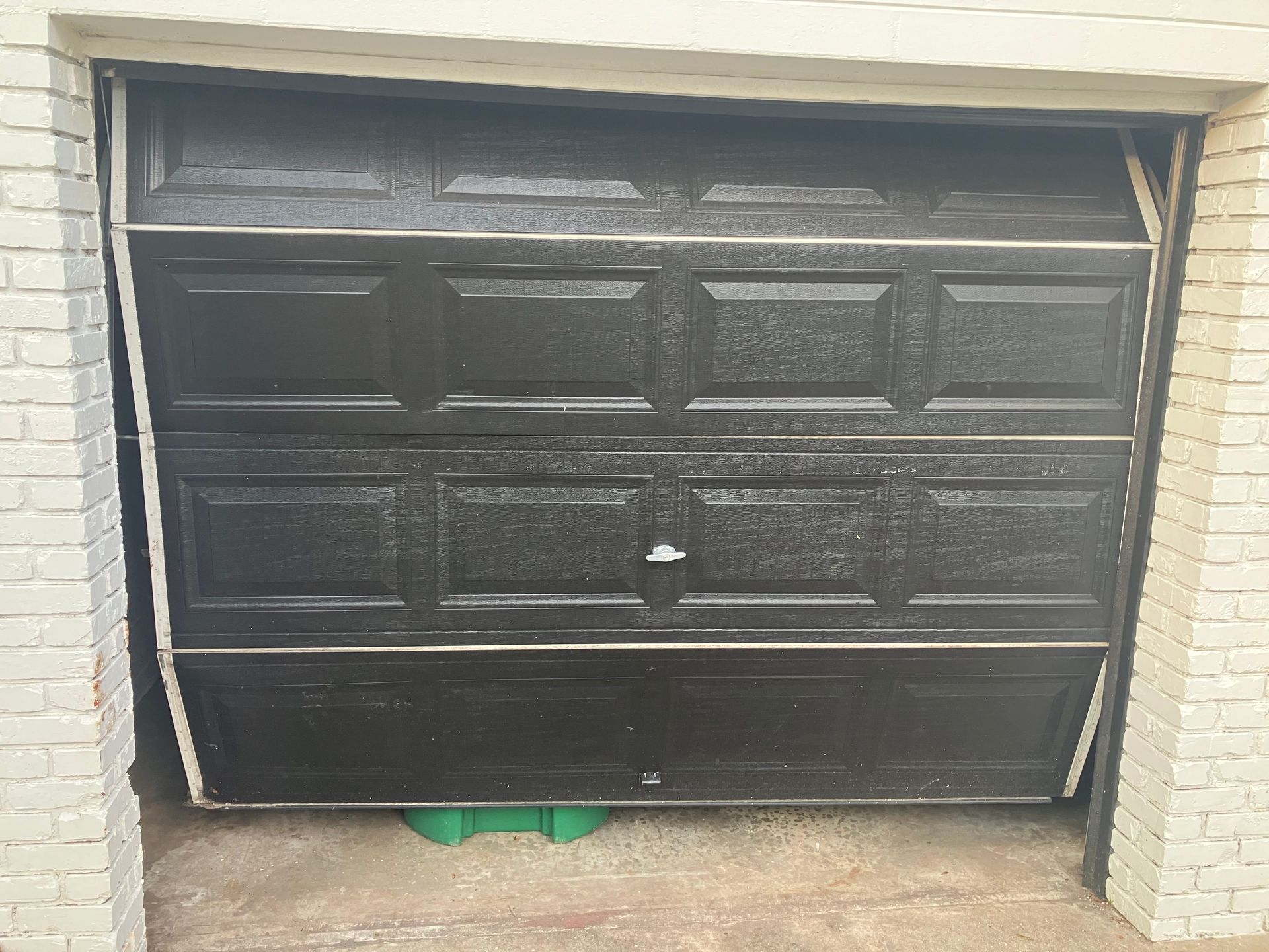 Black garage door stuck slightly open with green bins visible underneath