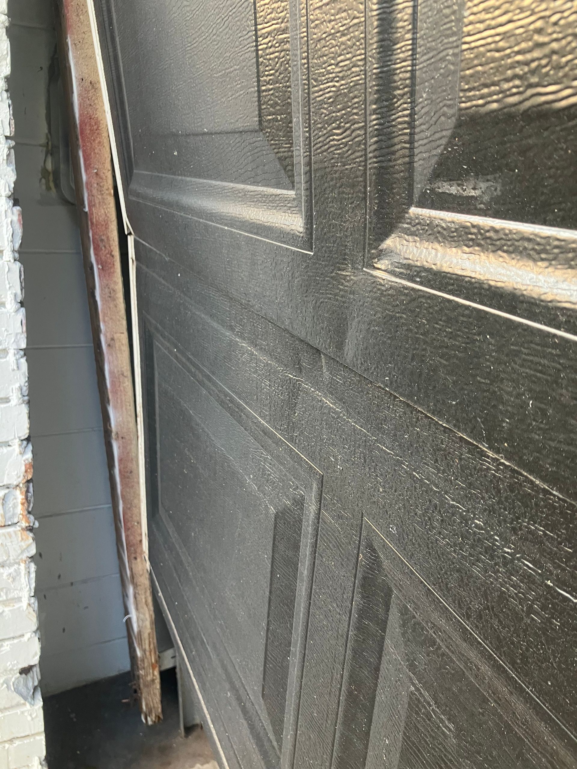 Black garage door slightly open beside a brick wall, showing a narrow gap.