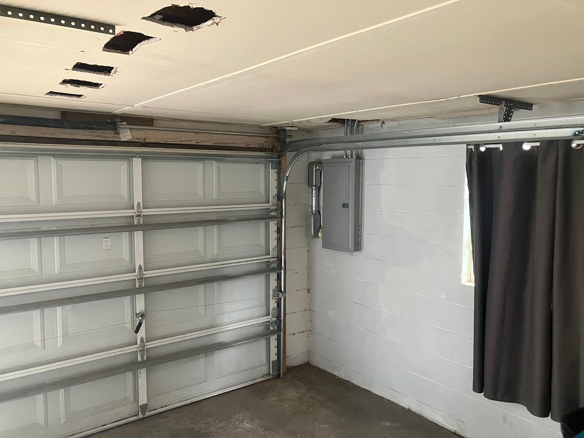 Empty garage interior with metal shelving, exposed ceiling, electrical panel, and a curtained window