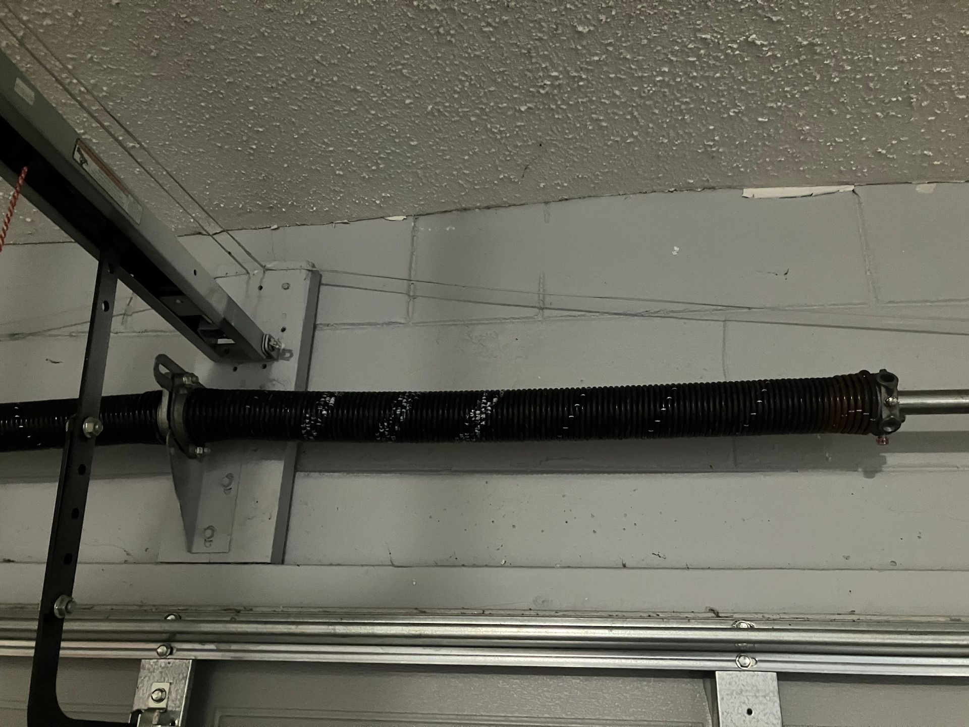 Black coiled garage door torsion spring mounted above a white door track under a speckled ceiling