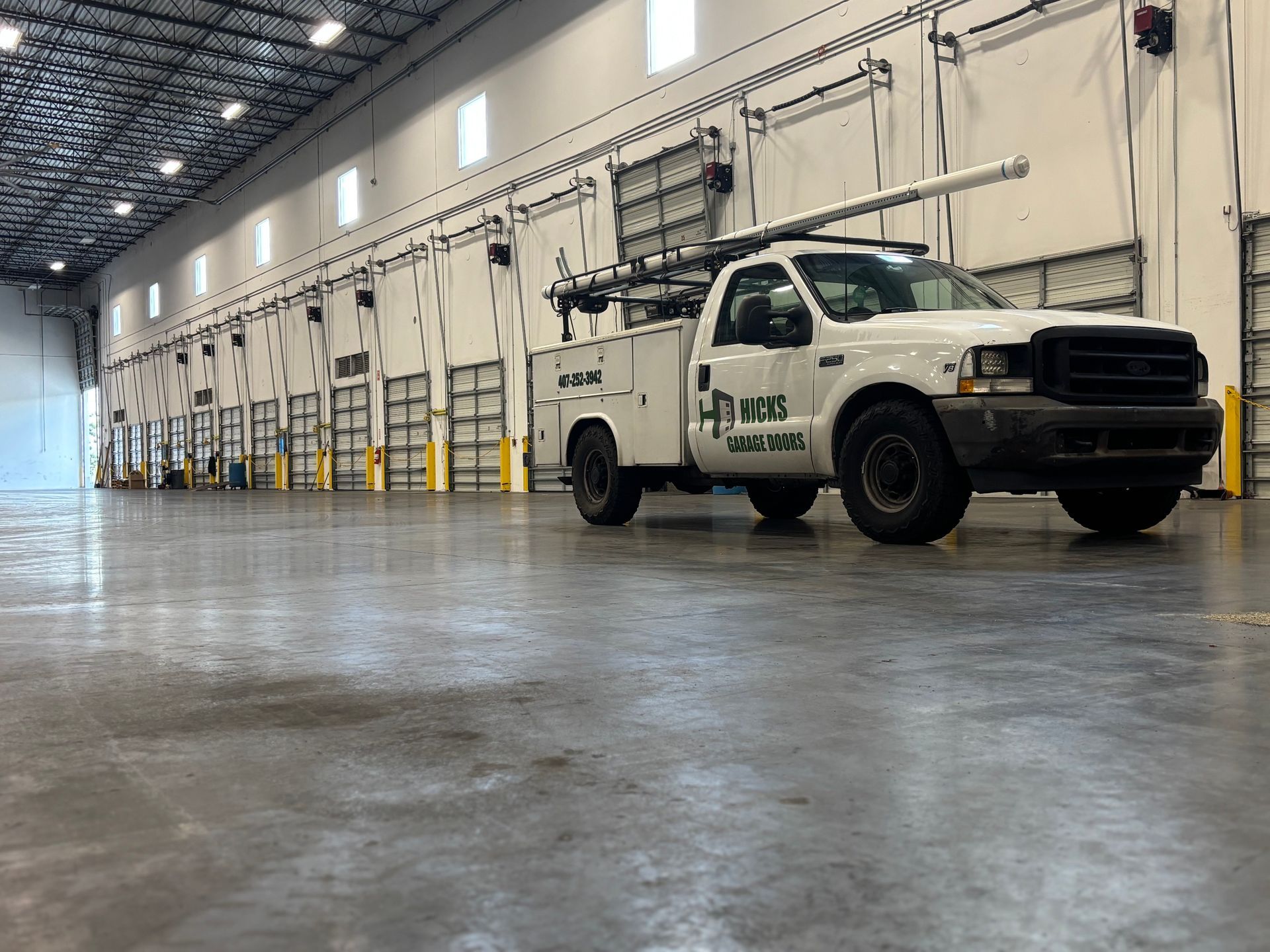 White utility pickup truck parked inside a large industrial warehouse with garage doors and bright overhead lights.