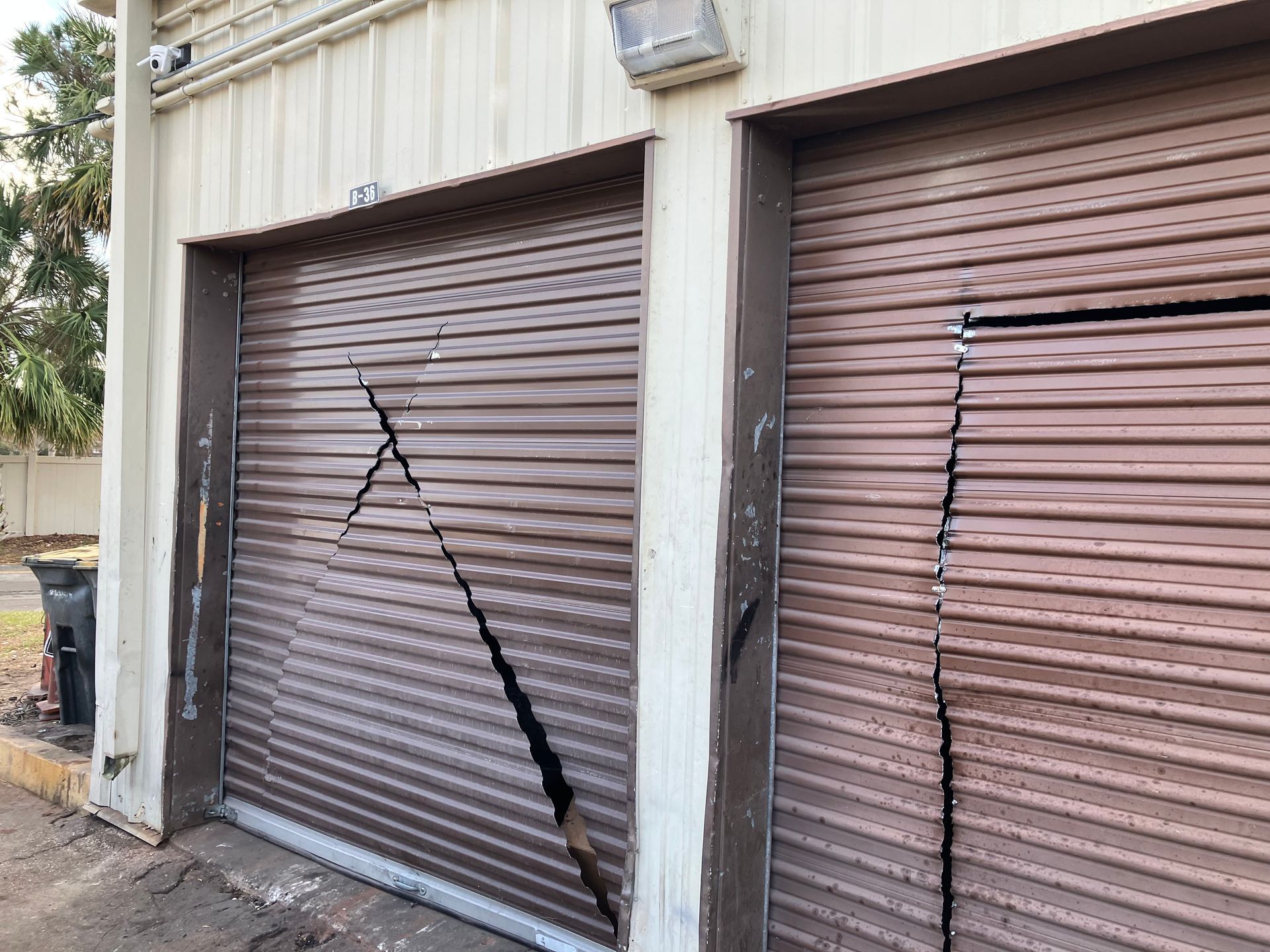 Damaged metal roll-up garage doors with a large crack in one door and a beige building exterior.