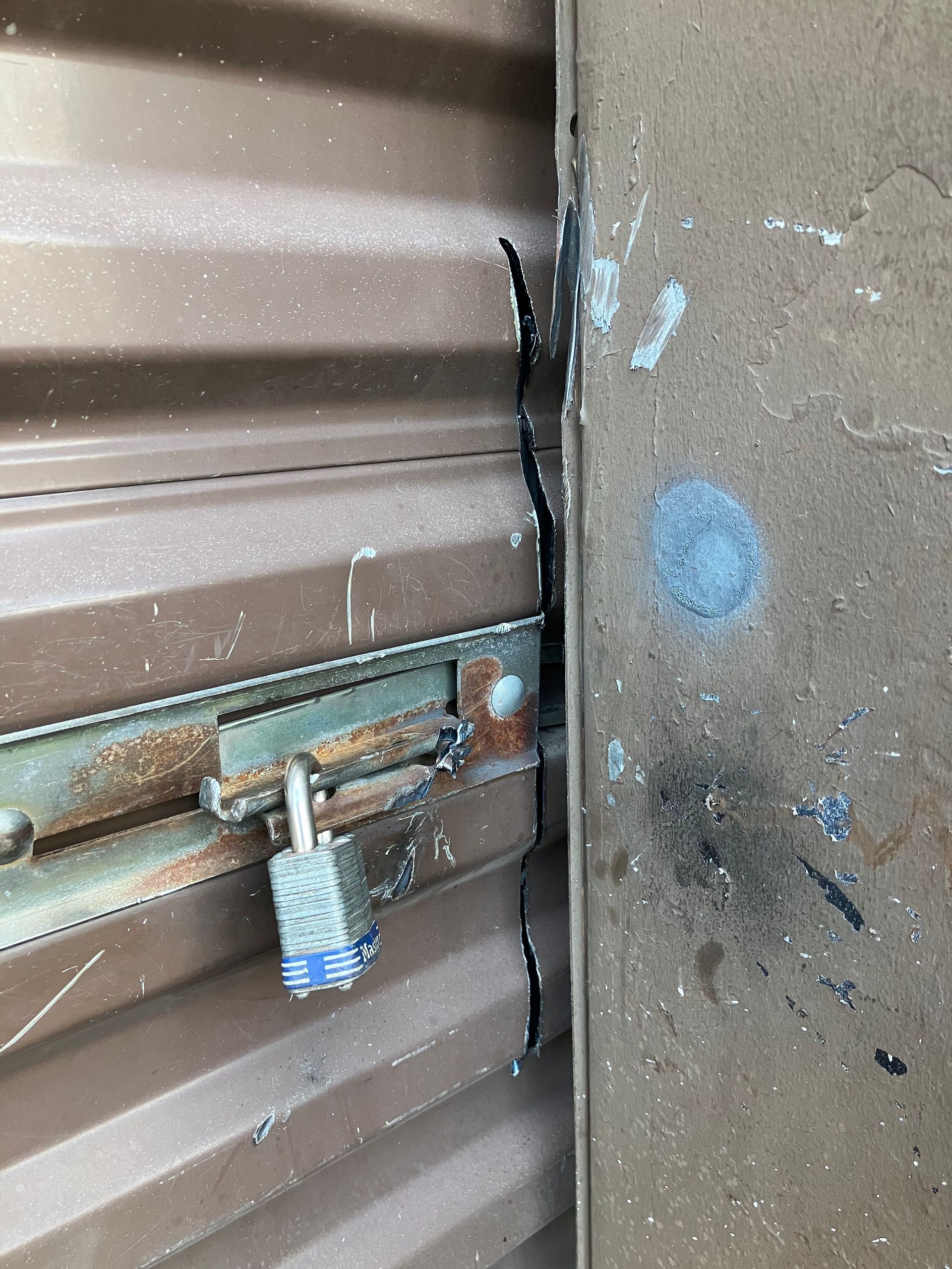 Close-up of a rusty latch and padlock securing a brown metal roll-up door against a concrete wall.