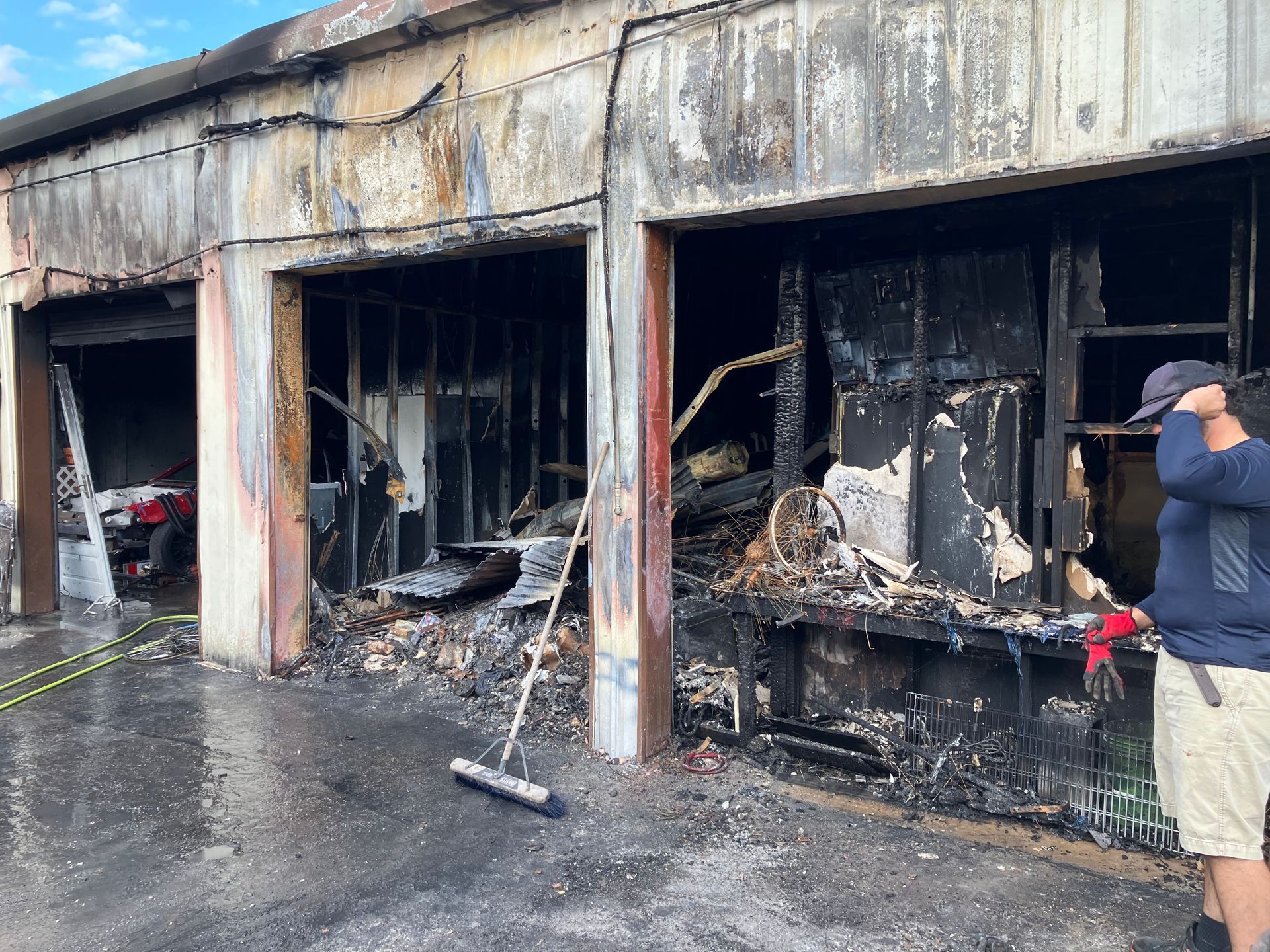 Burned-out garage bays with fire damage and charred debris; wet pavement and a person standing at right