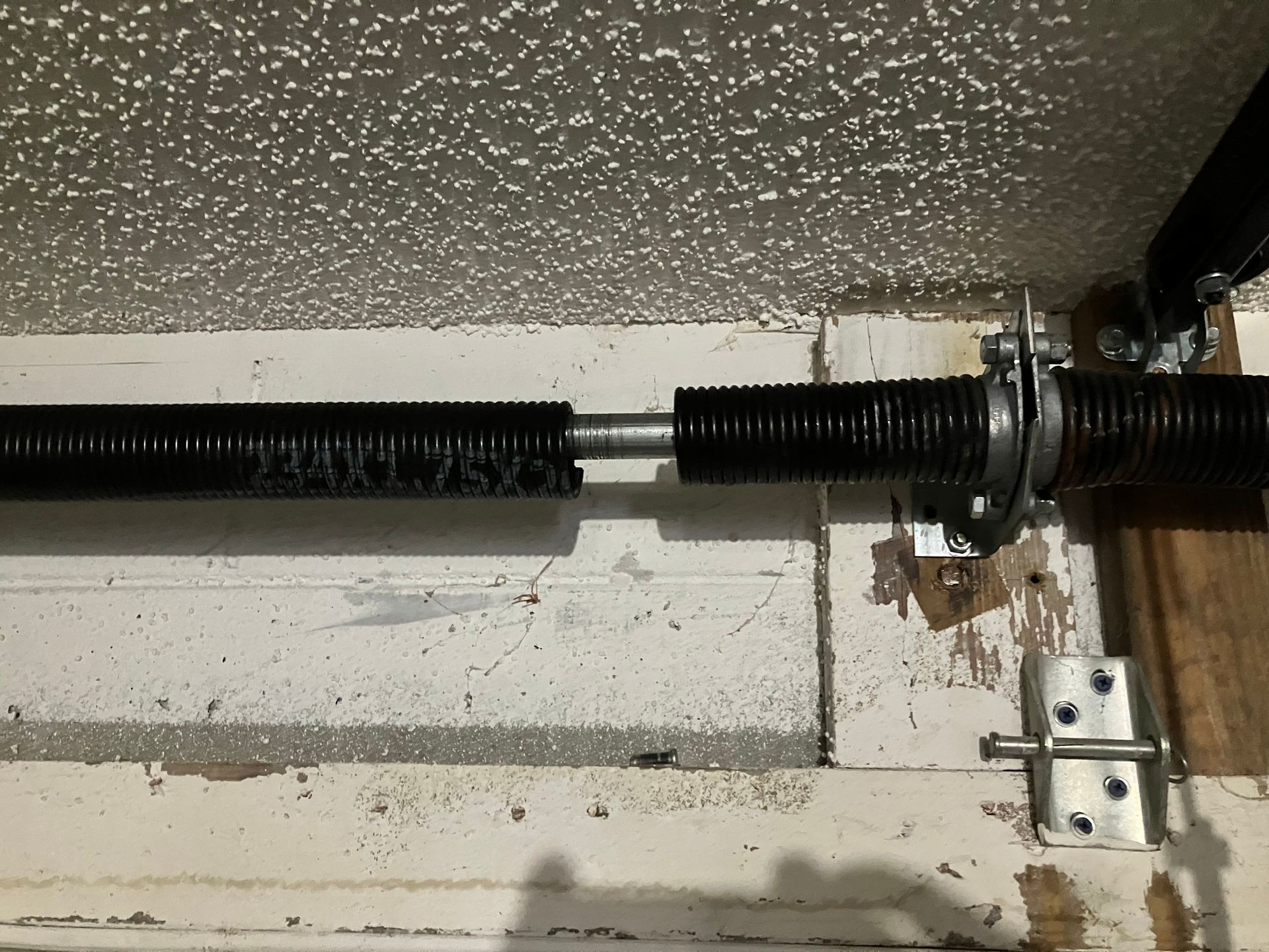 Black garage door torsion spring and metal hardware mounted above a white wall.