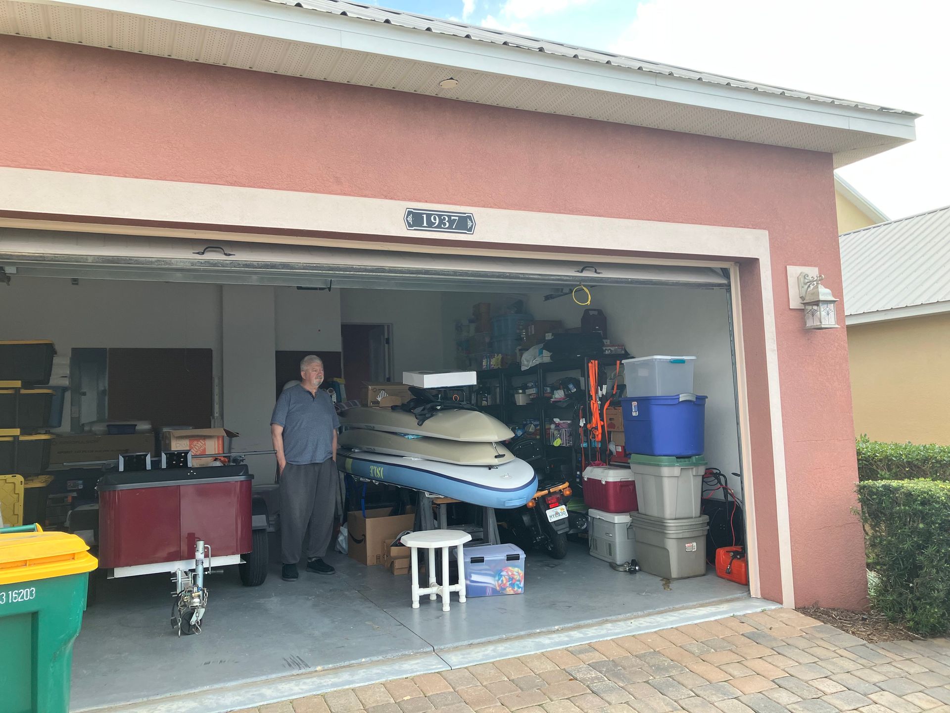 Open garage with man standing by stacked jetskis, tools, and storage bins inside a pink house.