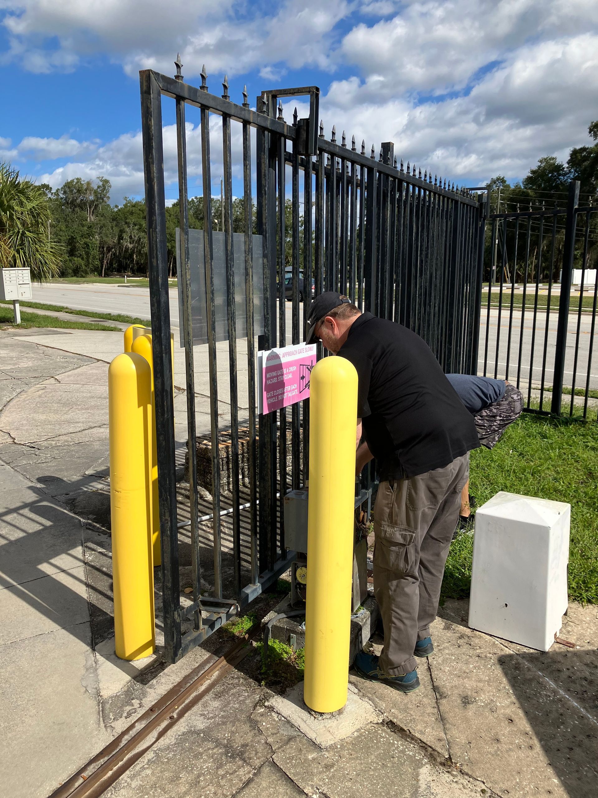 Man opening a black metal gate beside yellow bollards and a pink sign