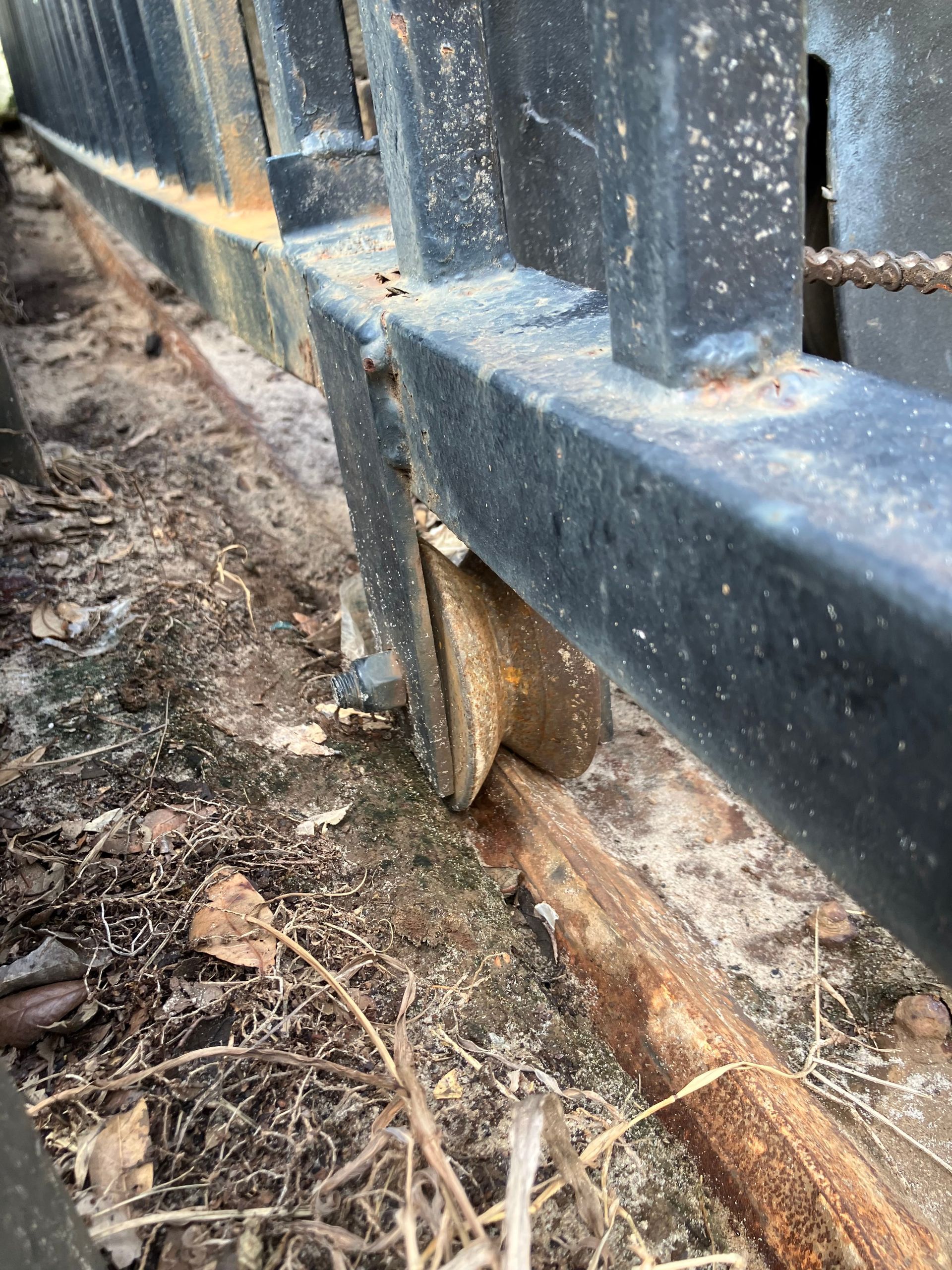 Close-up of a black metal fence post base set in dirt beside a wooden beam and gravel.