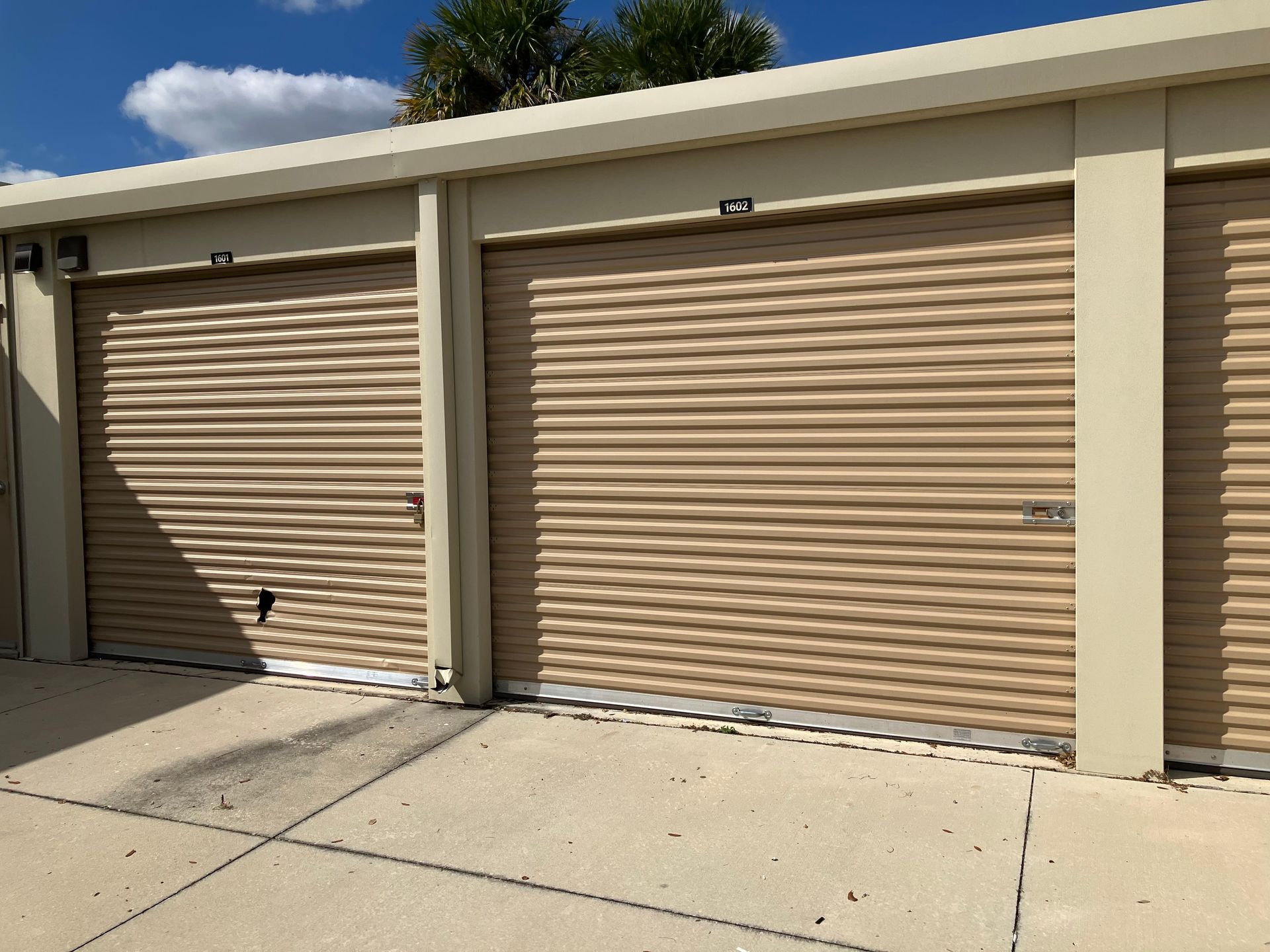 Two beige roll-up garage doors in a storage unit row under a blue sky.