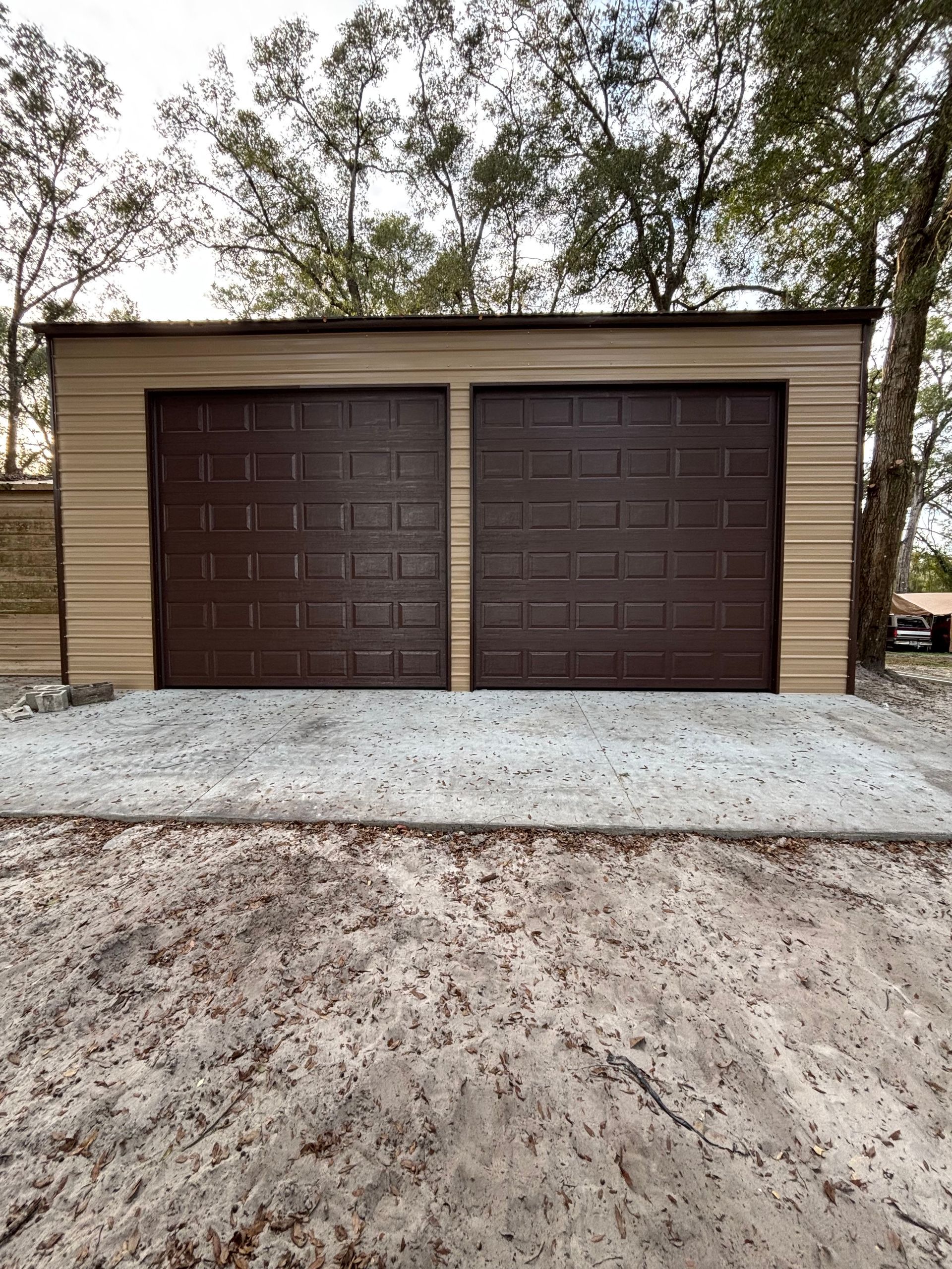 Two brown garage doors on a beige detached garage with a gravel driveway and trees in the background