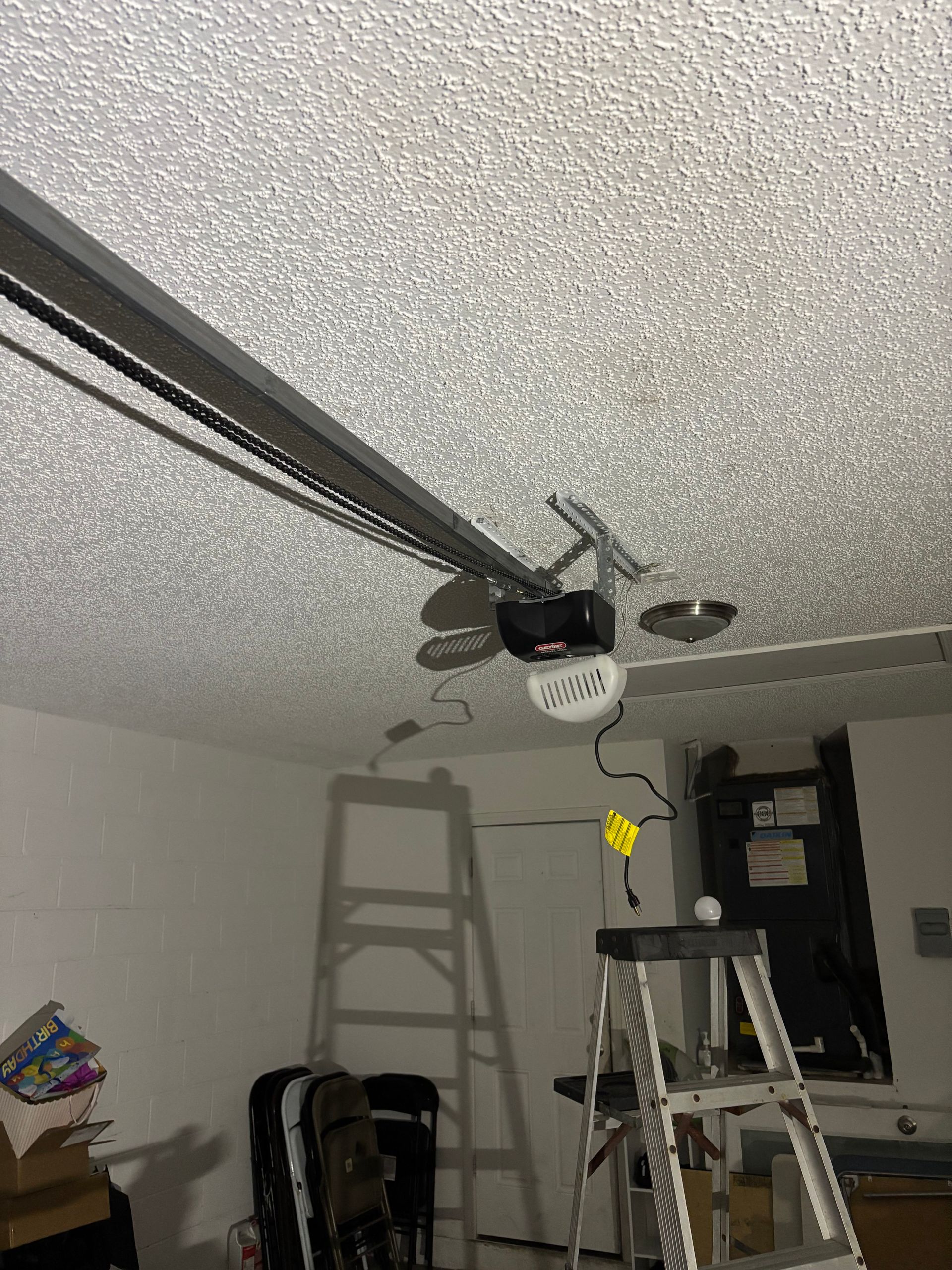 Garage ceiling with a ladder beneath a ceiling fan and track rails, near a window and storage items.