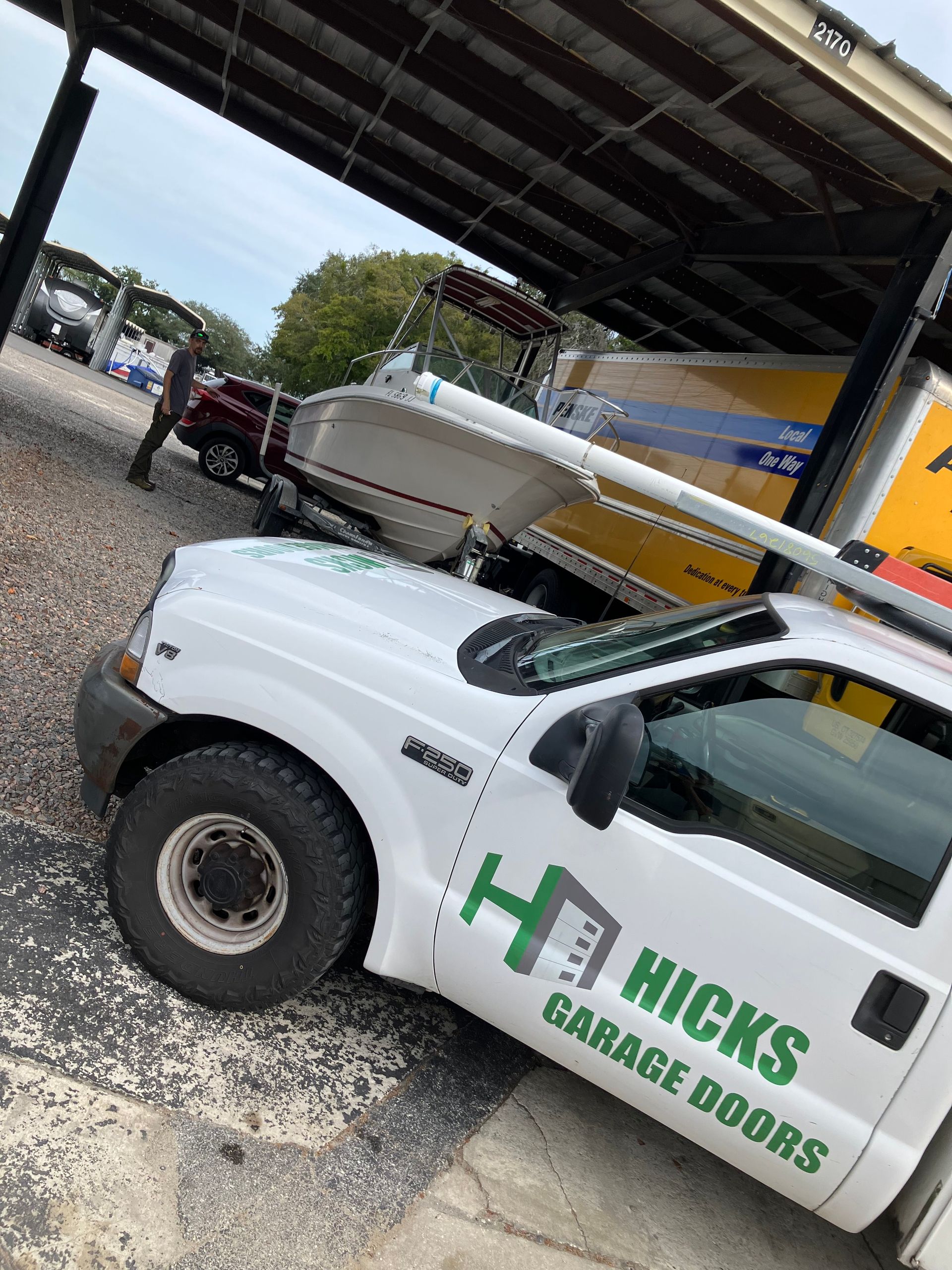 White Hicks Garage Doors service truck under a covered area beside a trailer and gravel lot