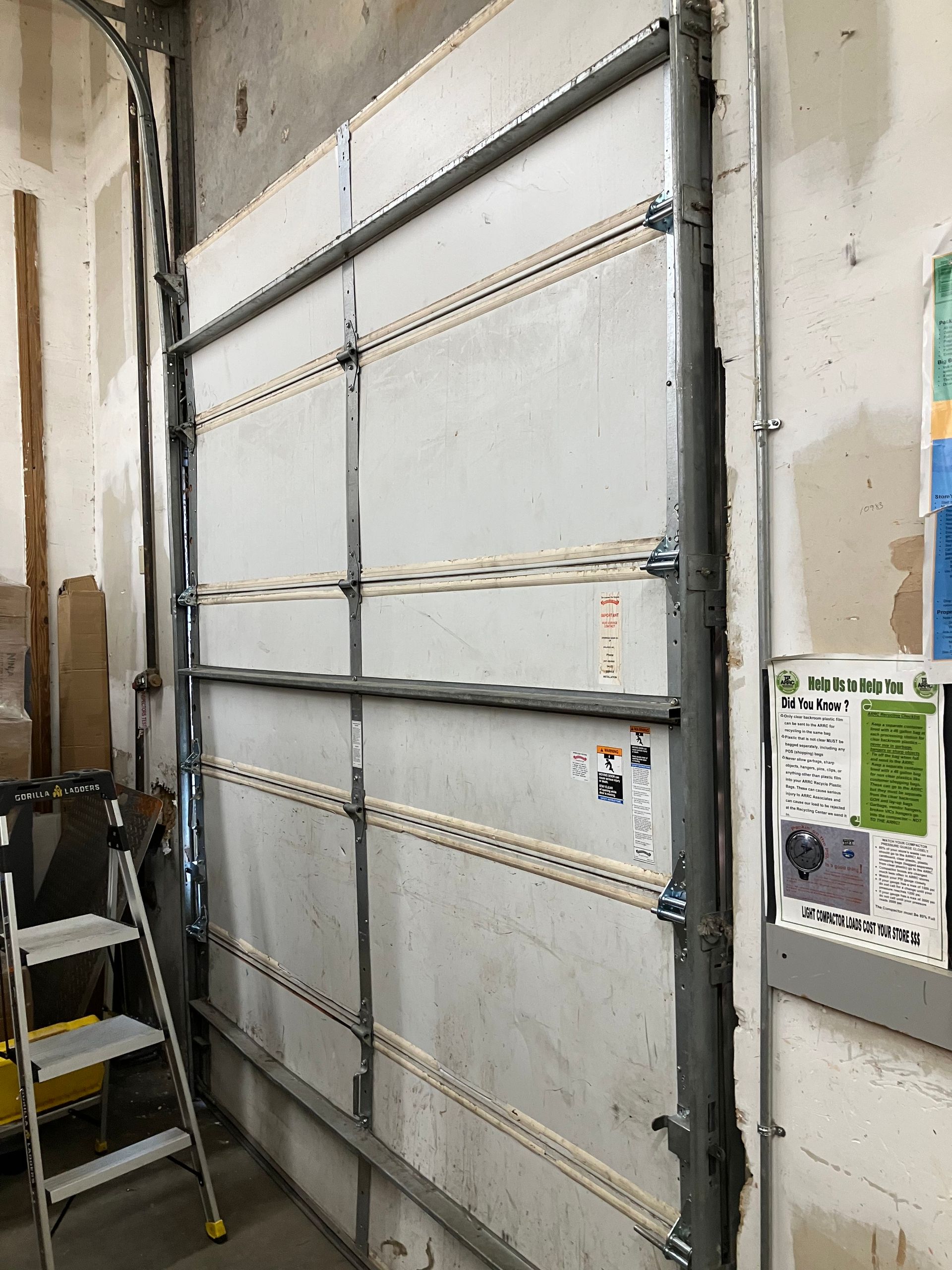 Large gray industrial garage door in a workshop, with a step ladder beside it and wall-mounted controls on the right.