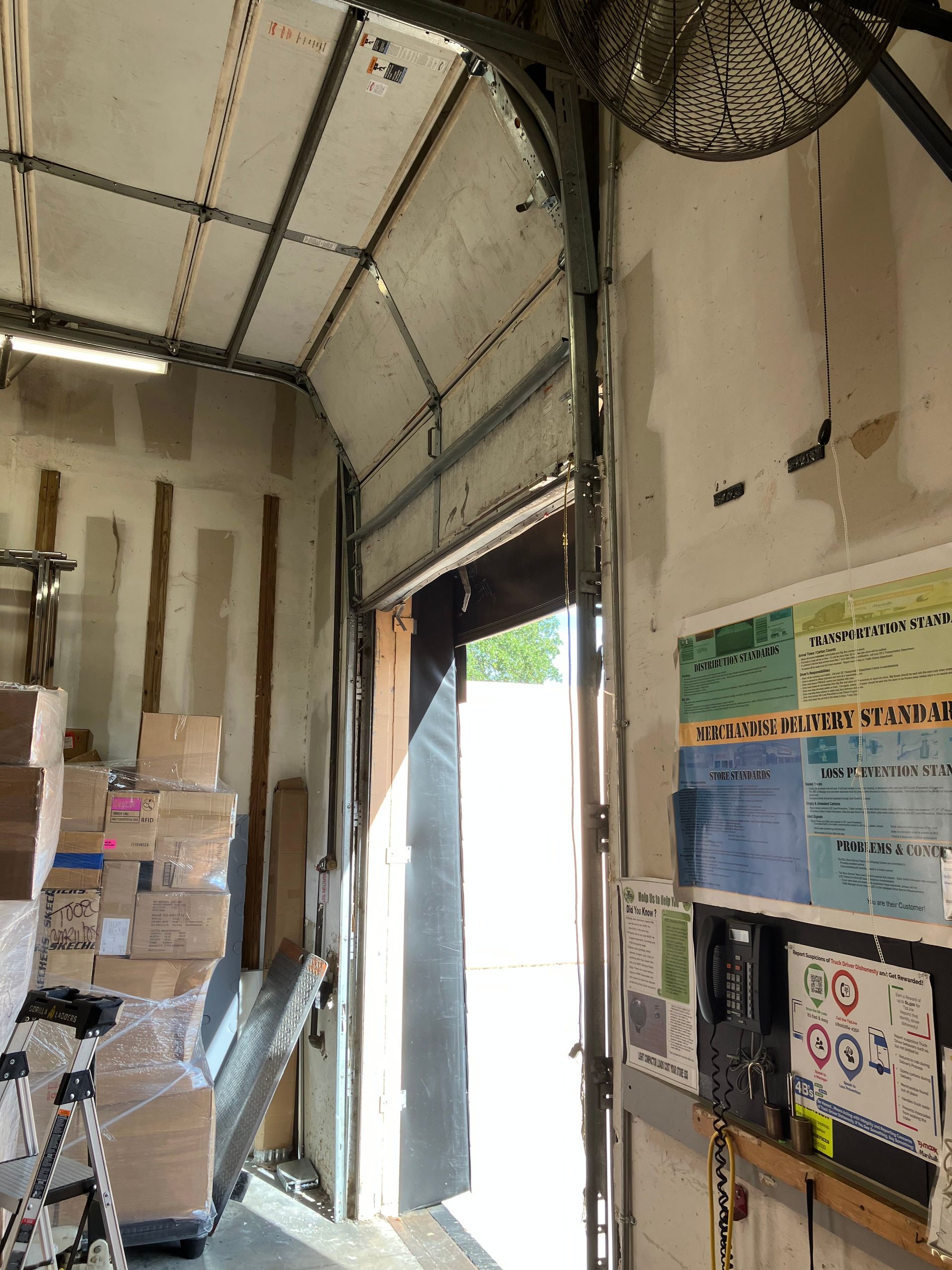 Narrow warehouse hallway with stacked boxes, open door to bright light, and safety posters on the wall