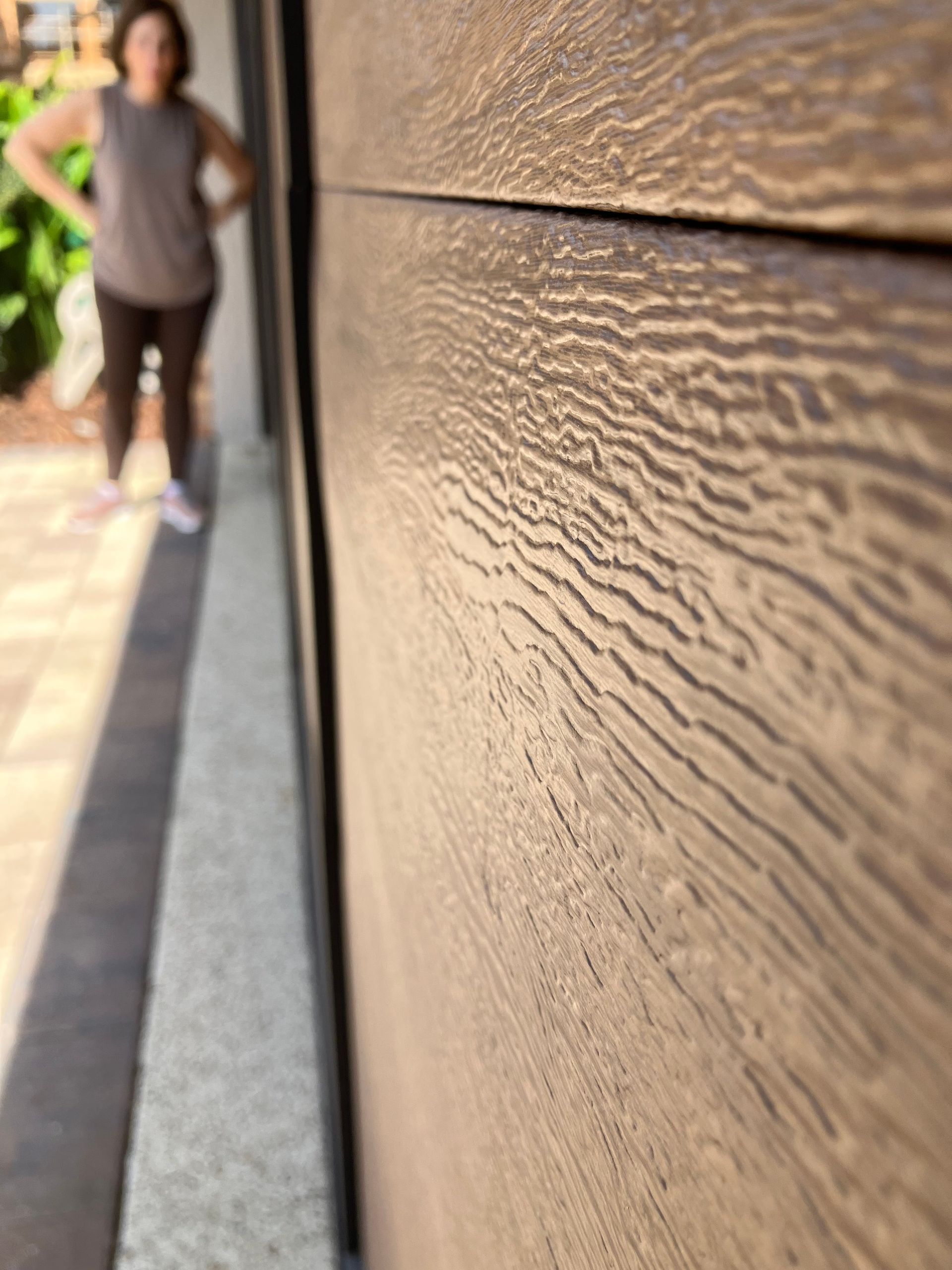 Close-up of textured wooden wall beside a sidewalk, with a blurred person walking in the background.