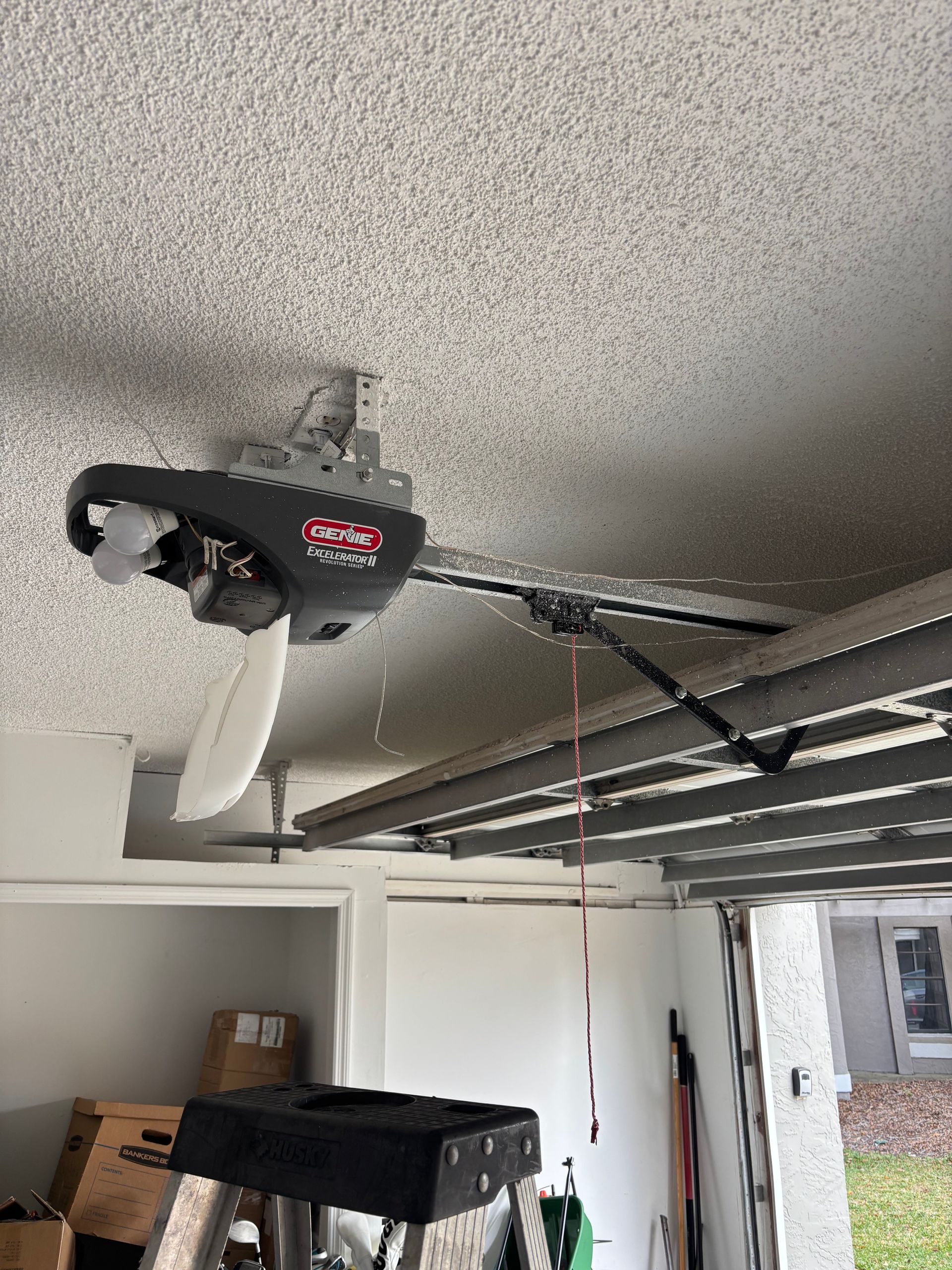 Ceiling-mounted garage door opener with chain rail and red emergency cord in a garage.