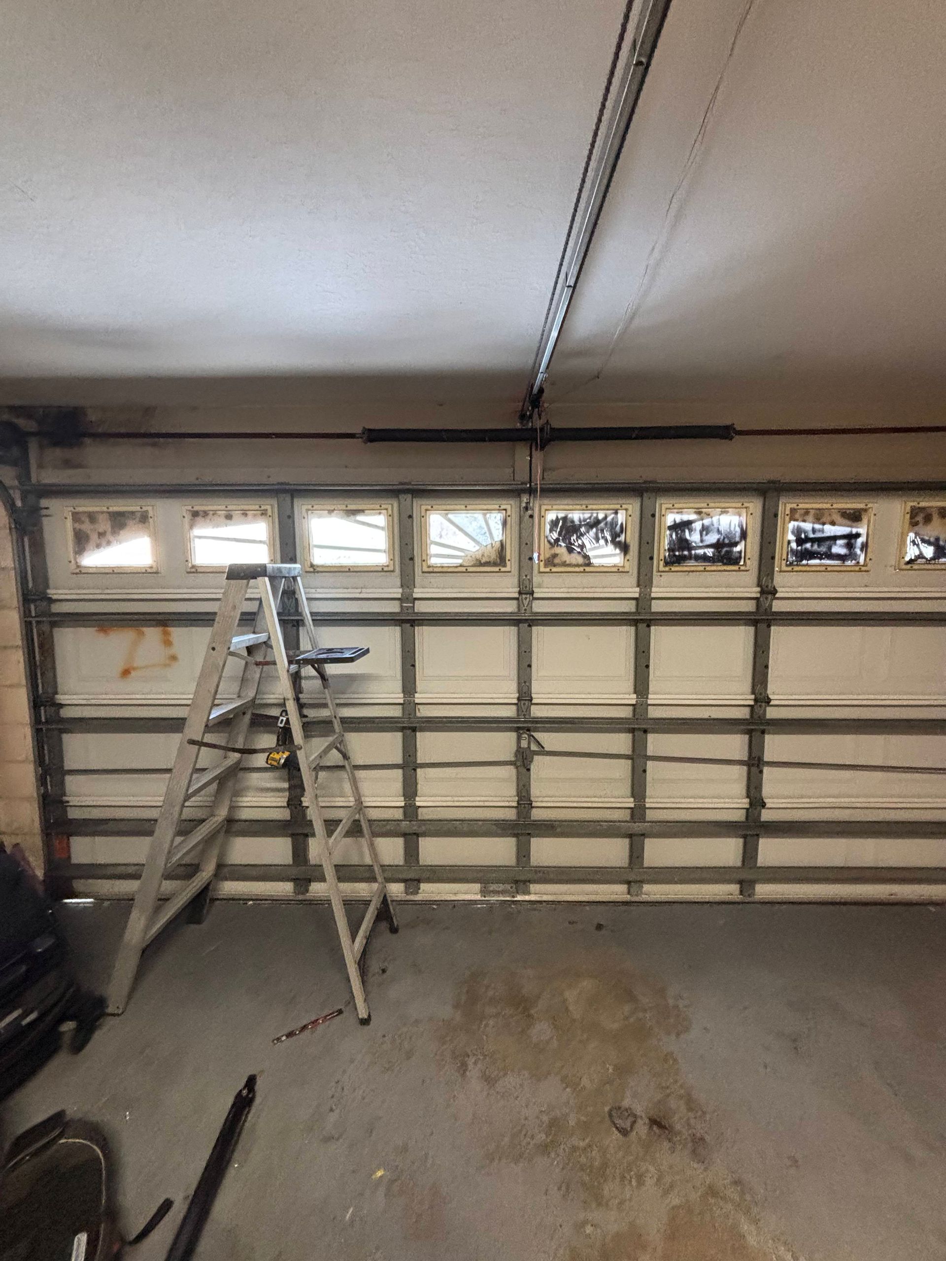 Ladder in a garage beside a closed garage door, with storage items and a stained concrete floor.