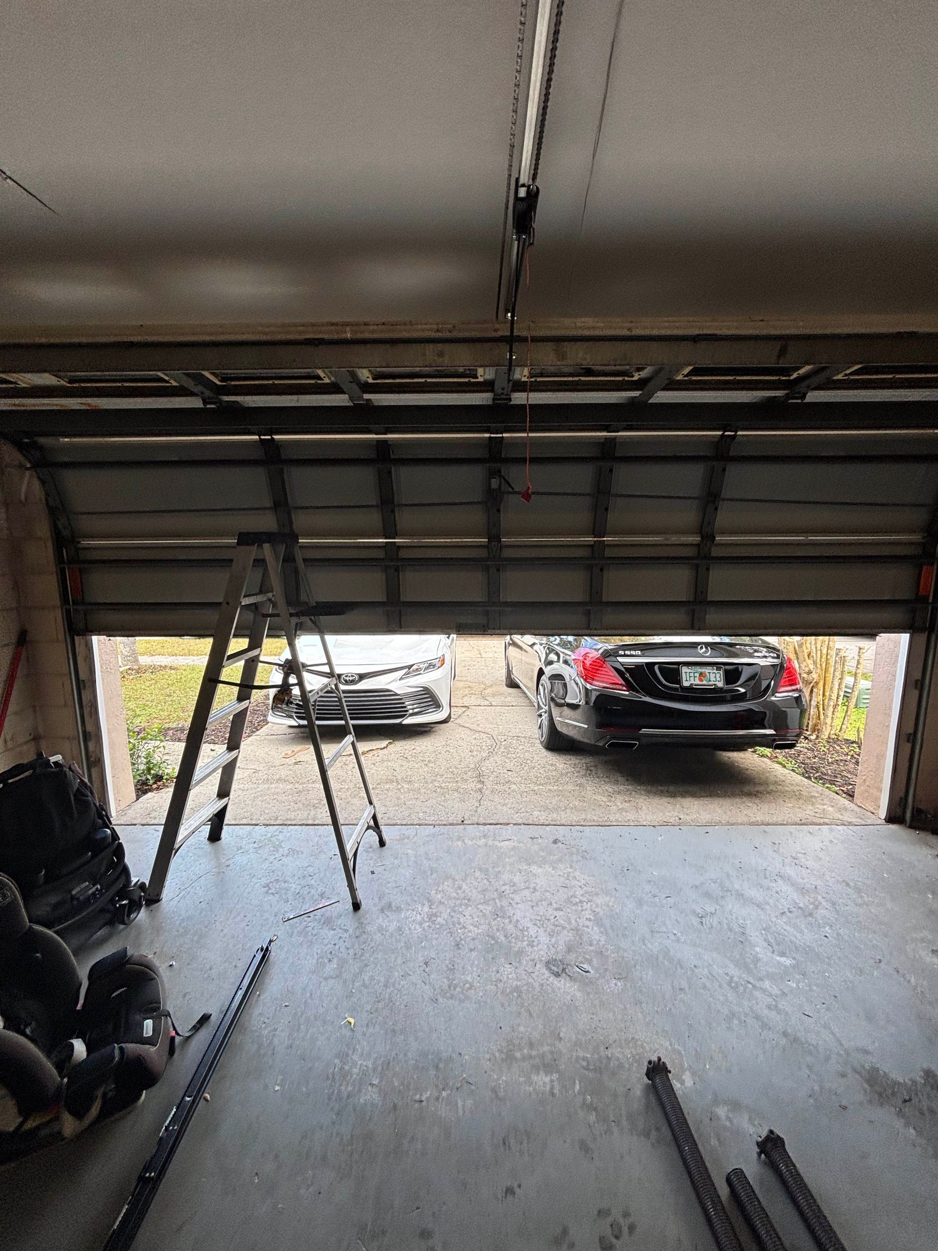 Open garage with a gray car parked outside on a gravel driveway and a black car near the driveway exit.