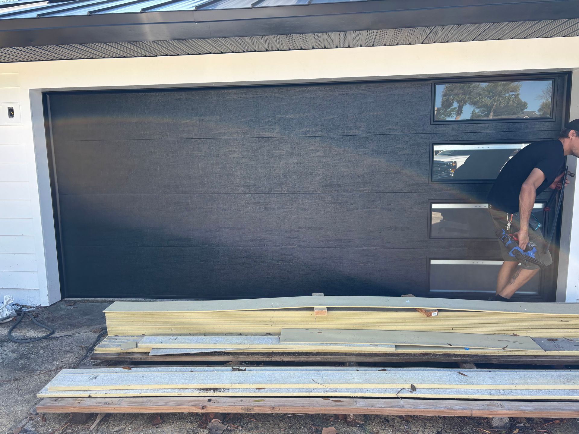 Black garage door with stacked wood planks in front and a person partially visible at right