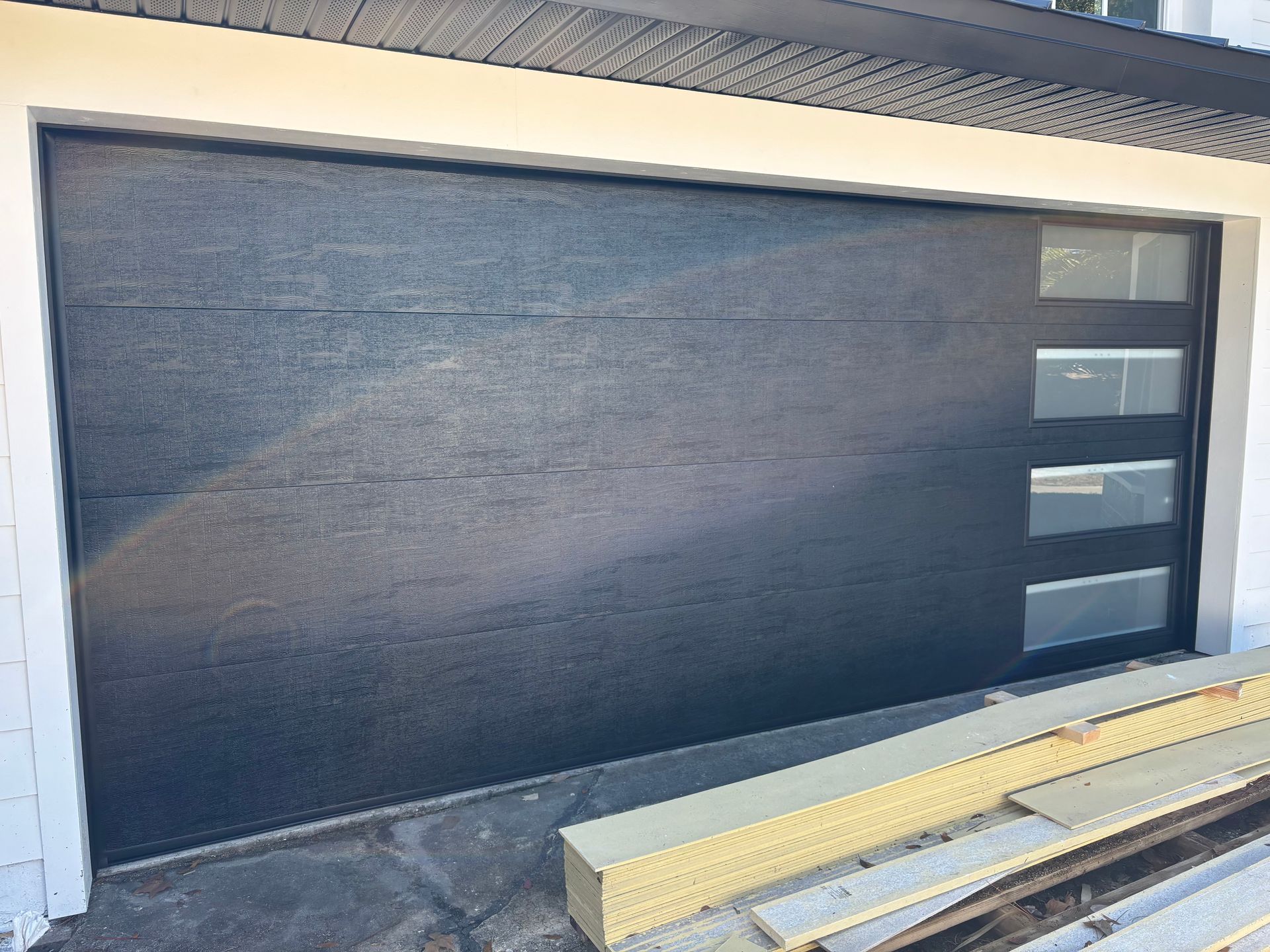 Black garage door with narrow windows on the right and lumber stacked in front.