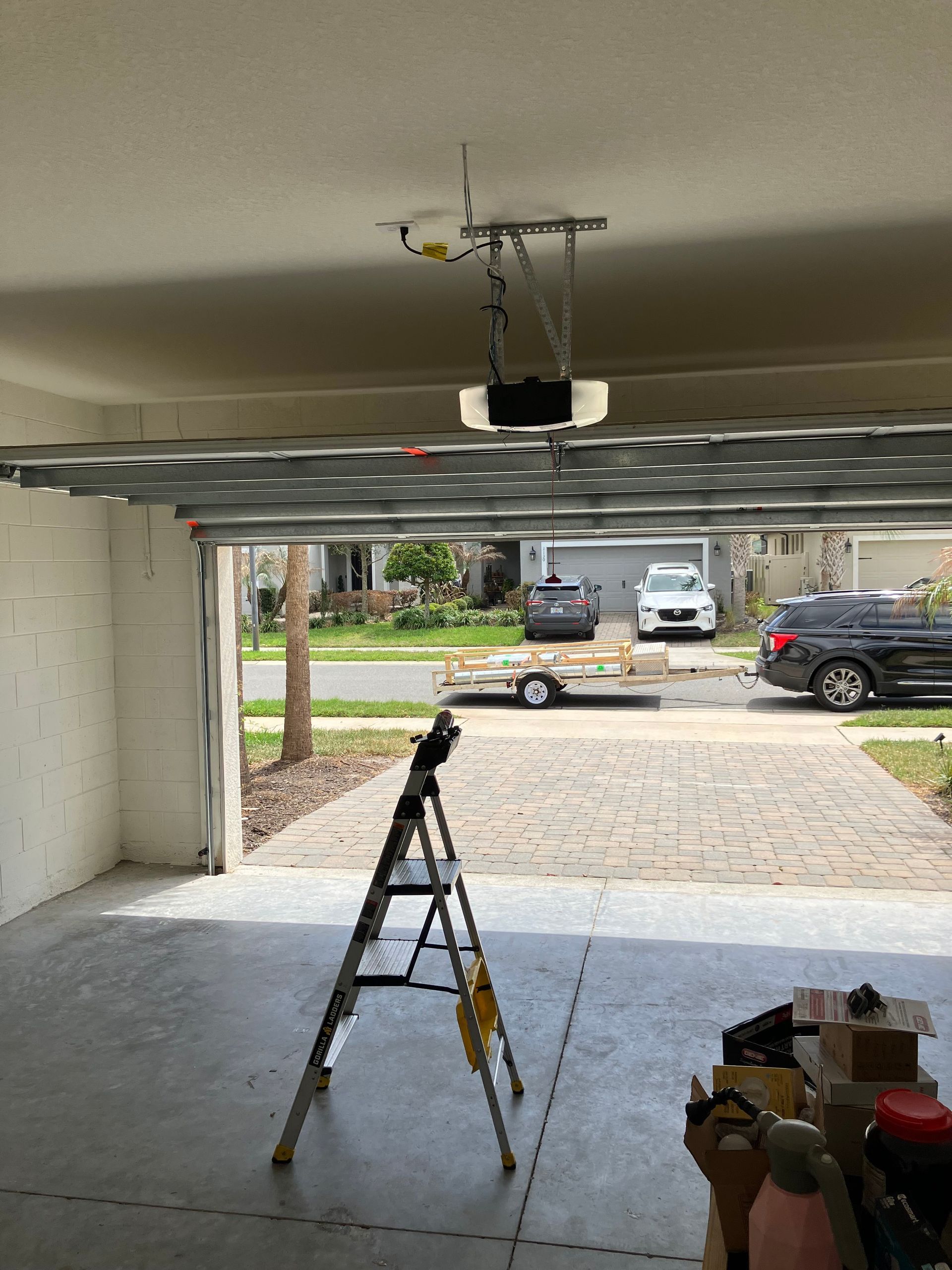 Garage interior with open door, step ladder, and tools on the floor, looking out to a driveway and parked vehicles
