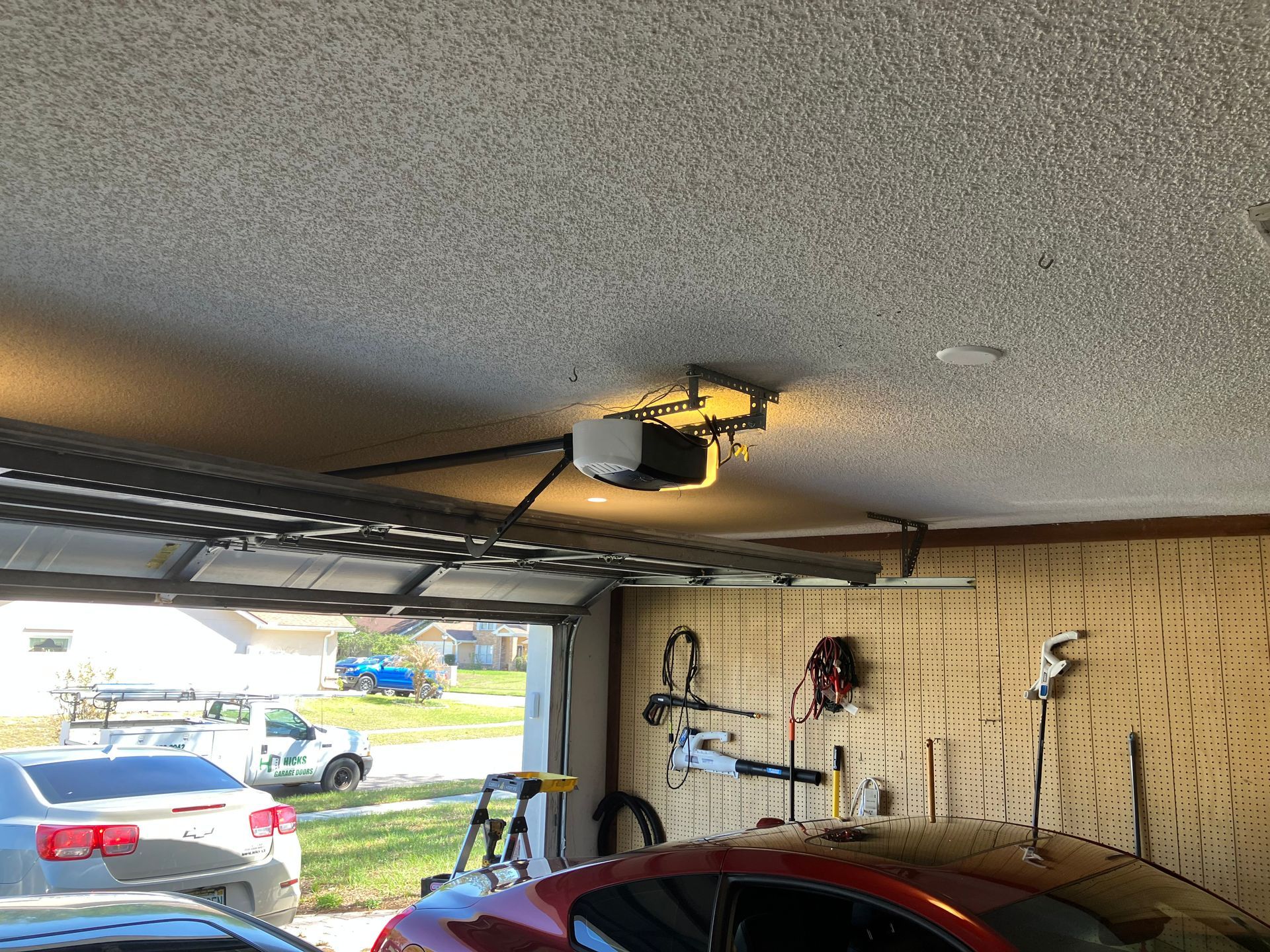 Garage interior with open door, parked car, and tools on the wall under a ceiling-mounted opener