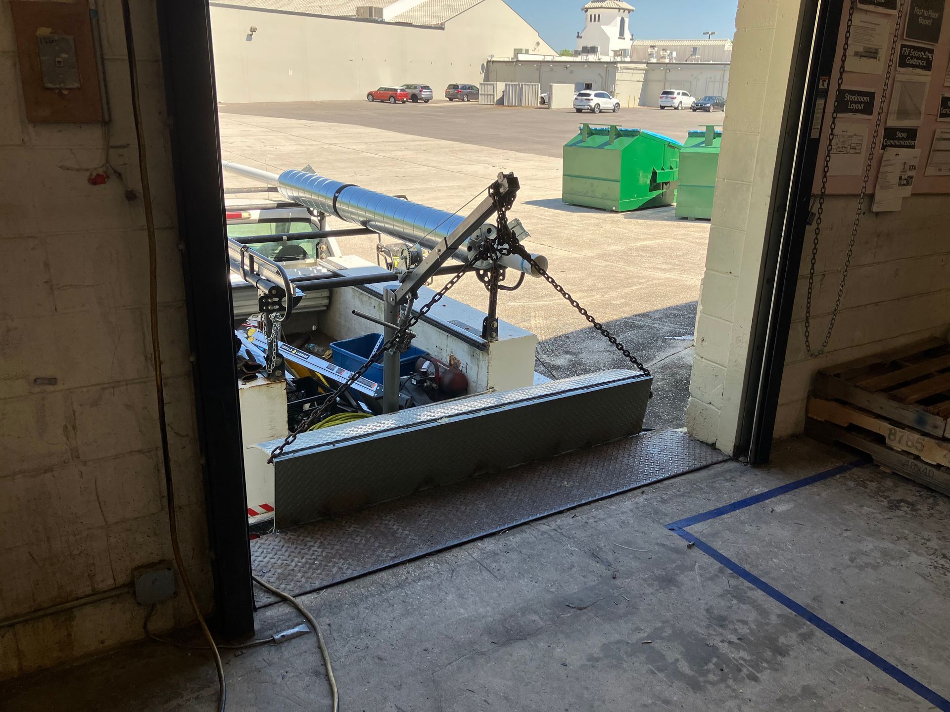 Wheelchair-accessible lawn mower at a building doorway, with outdoor pavement and a green dumpster beyond.