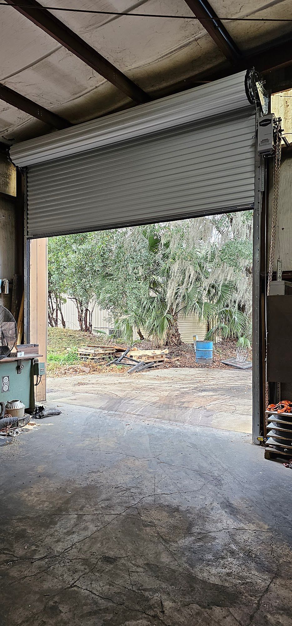Open garage with a partially raised shutter door, showing a dirt driveway and trees outside.