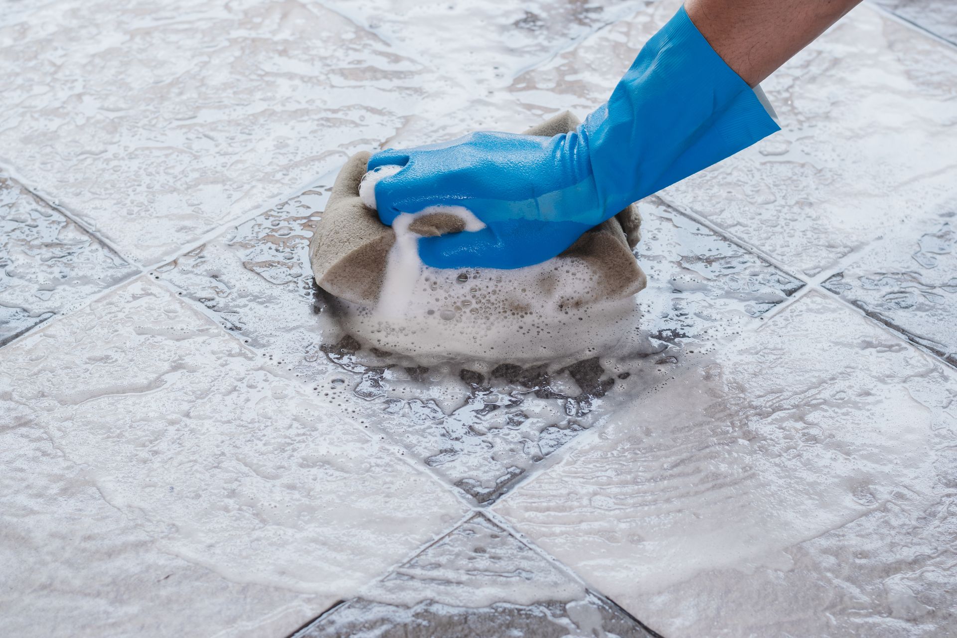 A person wearing blue gloves is cleaning a tile floor with a sponge.