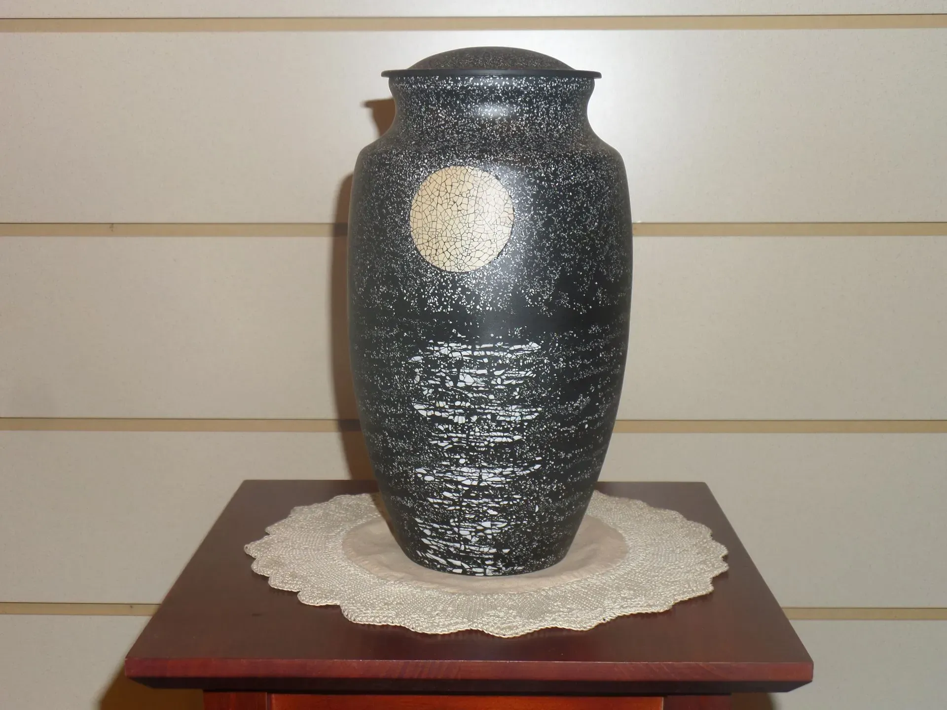 Black vase on a lace doily atop a small wooden table against a beige wall.
