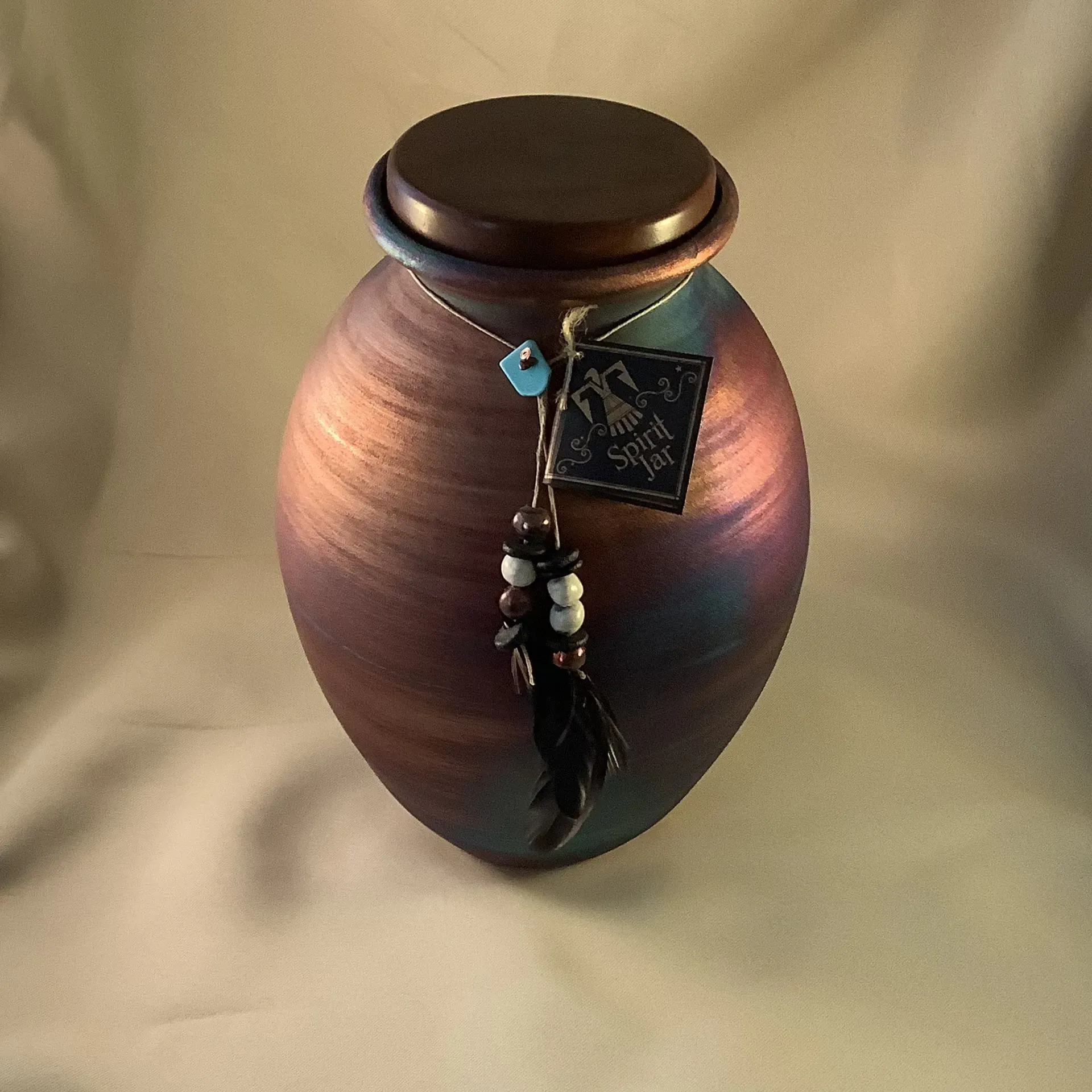 Decorative ceramic urn with a dark lid and bead-and-feather charm on a beige background